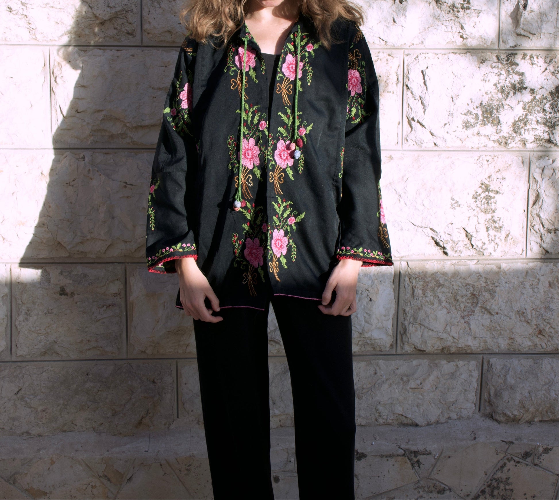 Woman wearing a black embroidered jacket with floral patterns against a stone wall.