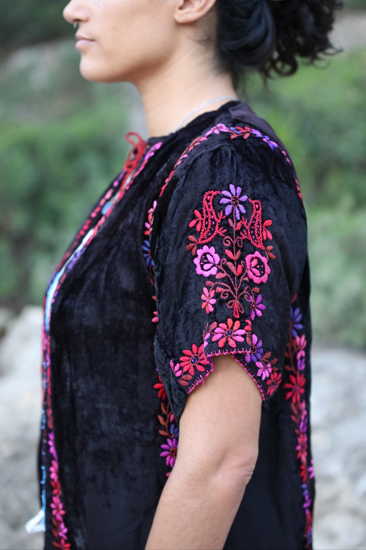 A woman shown in profile wearing a black embroidered top with pink, purple, and red floral patterns on the sleeves and neckline, photographed outdoors against a blurred natural background.