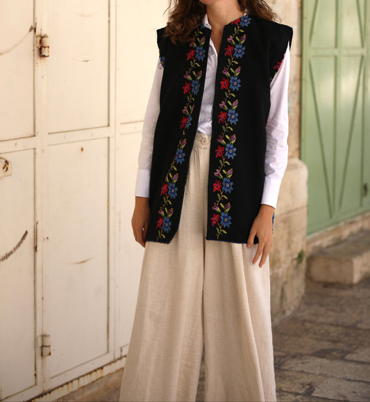 Woman wearing a black embroidered vest over a white shirt with beige pants, standing in front of a building.