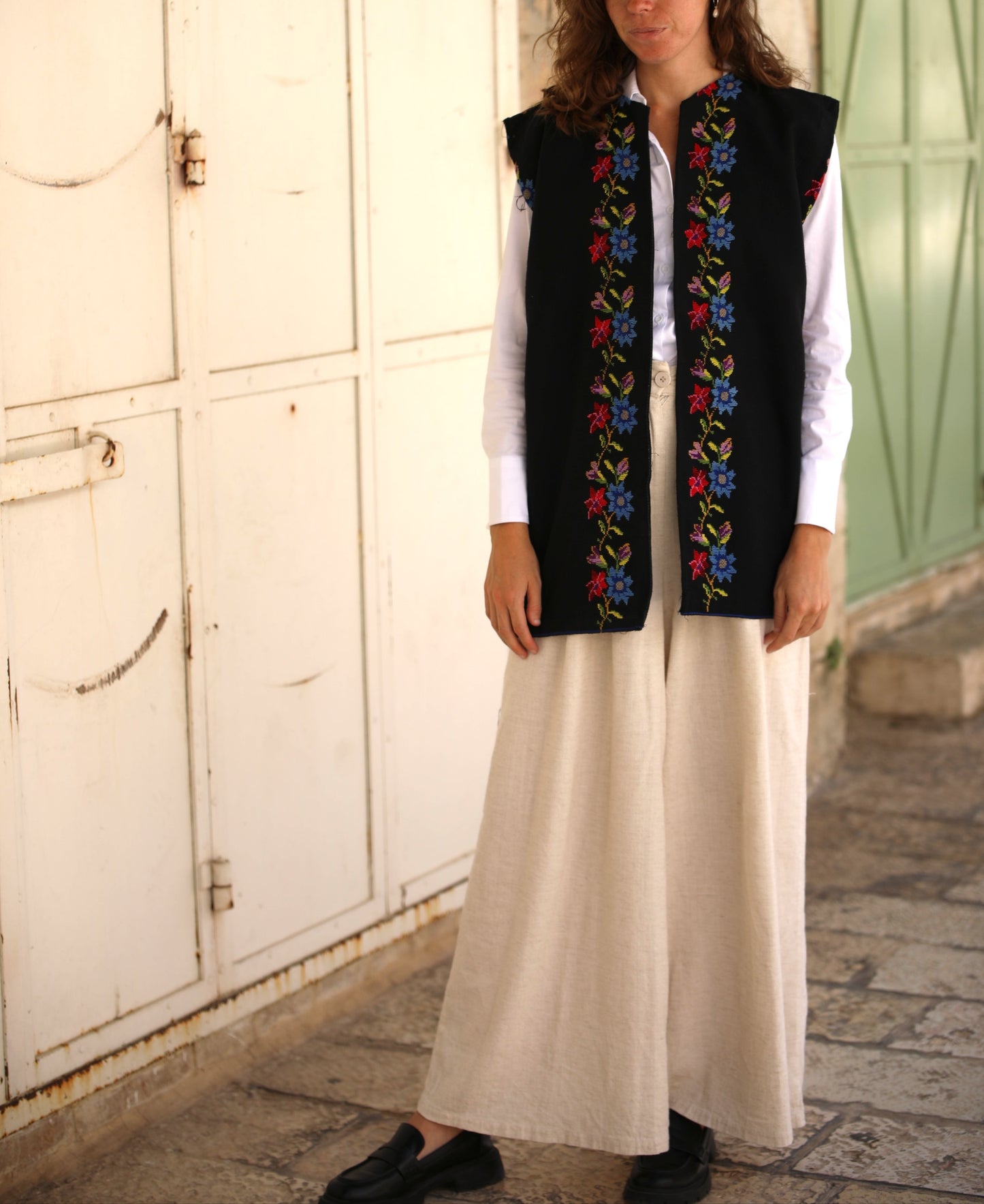 Woman wearing a black embroidered vest over a white shirt and beige pants, standing in front of a white wall.