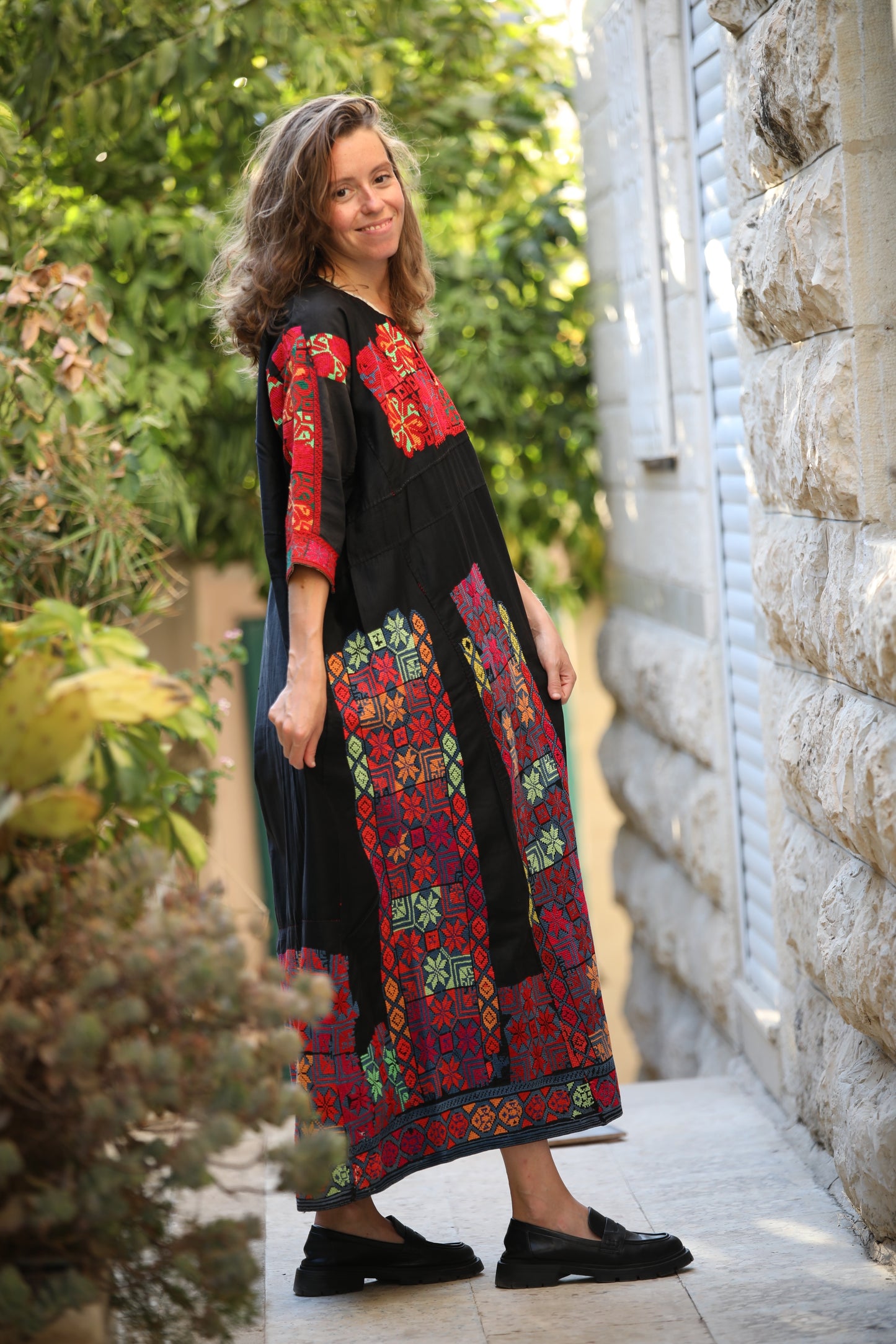 Woman wearing a colorful embroidered dress standing outdoors near a stone wall.