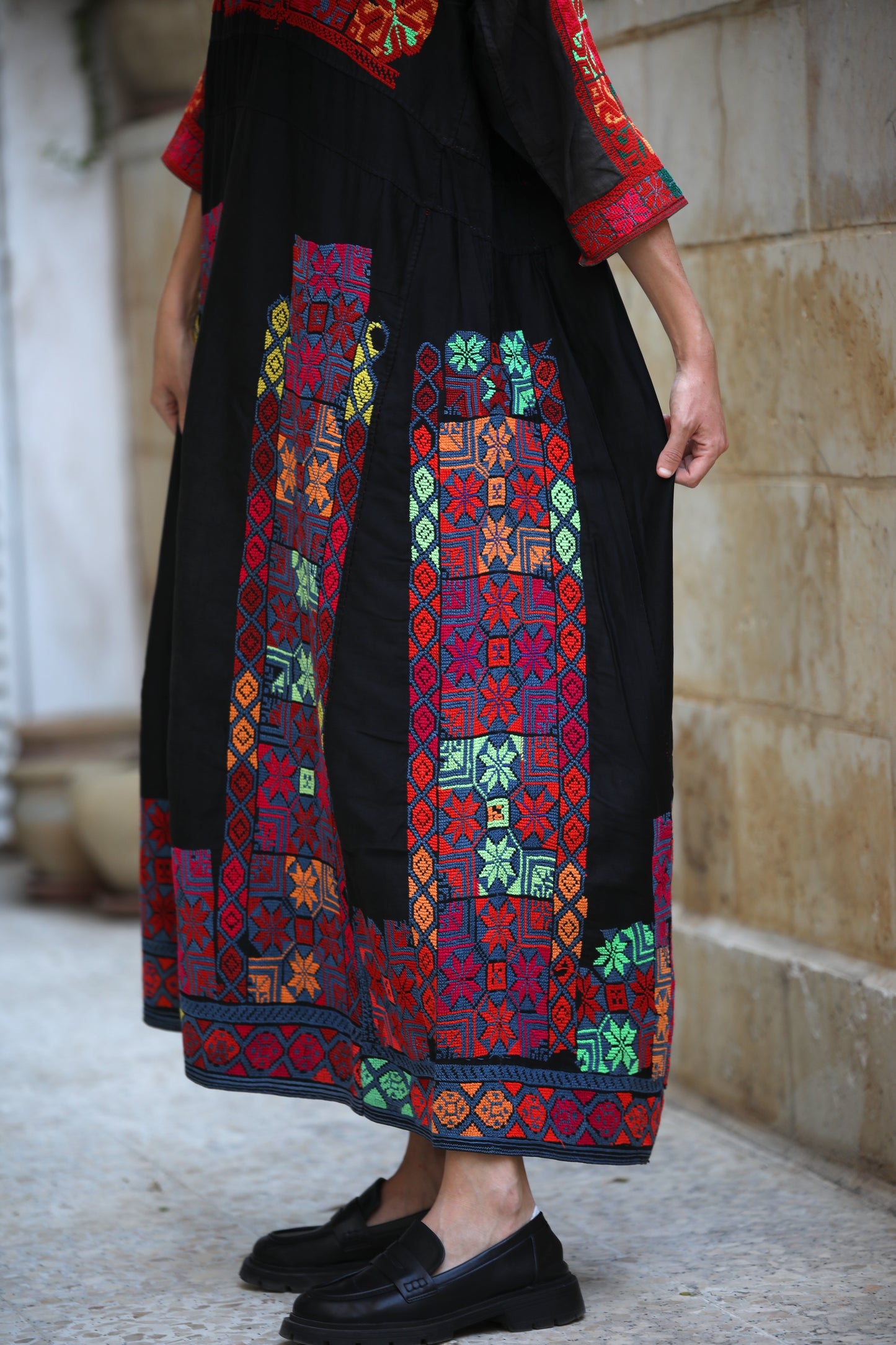Person wearing a black dress with colorful embroidery standing against a stone wall.