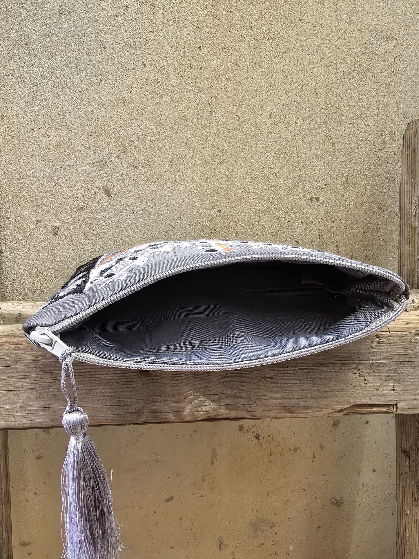 Gray clutch bag with embroidery and a tassel on a wooden surface.