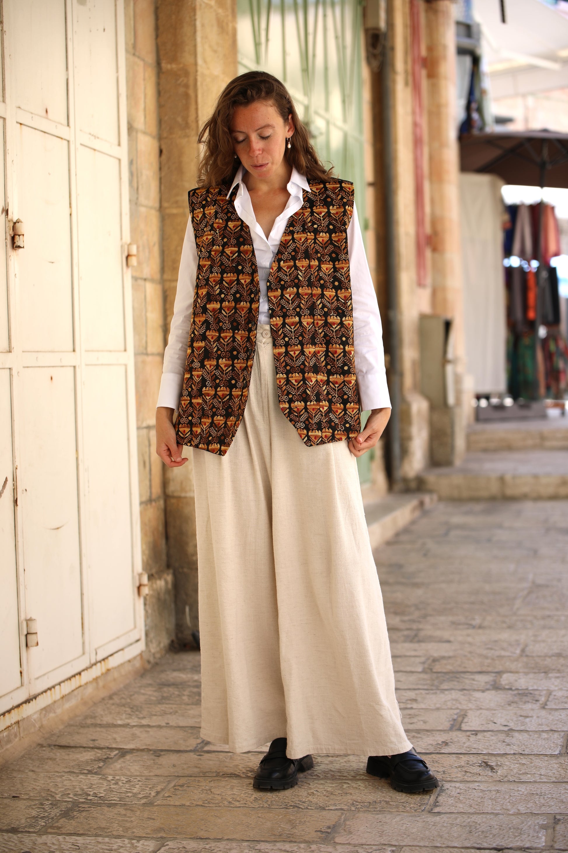 Woman wearing a patterned vest over a white shirt and beige pants on a street.
