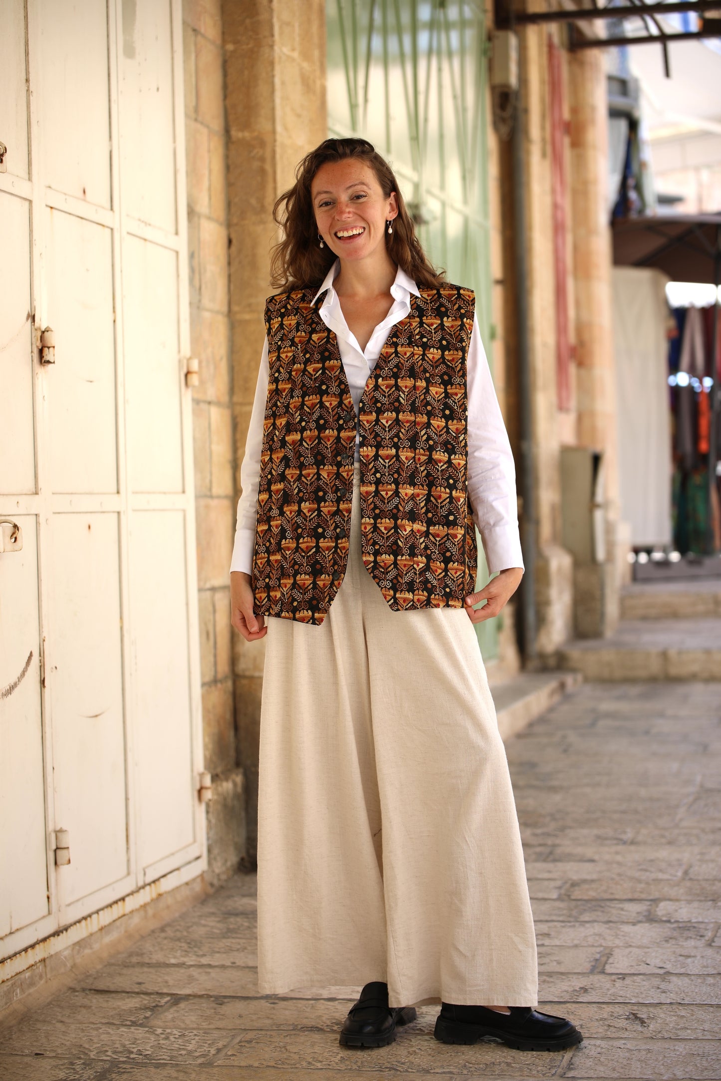 Woman wearing a patterned vest over a white shirt and beige pants, standing in an outdoor setting.