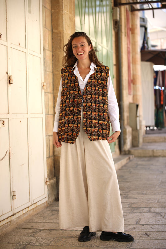 Woman wearing a patterned vest over a white shirt and beige pants, standing in an outdoor setting.