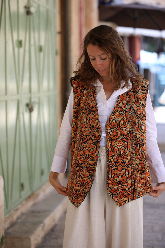 Woman wearing a patterned vest over a white shirt and beige pants on a street.