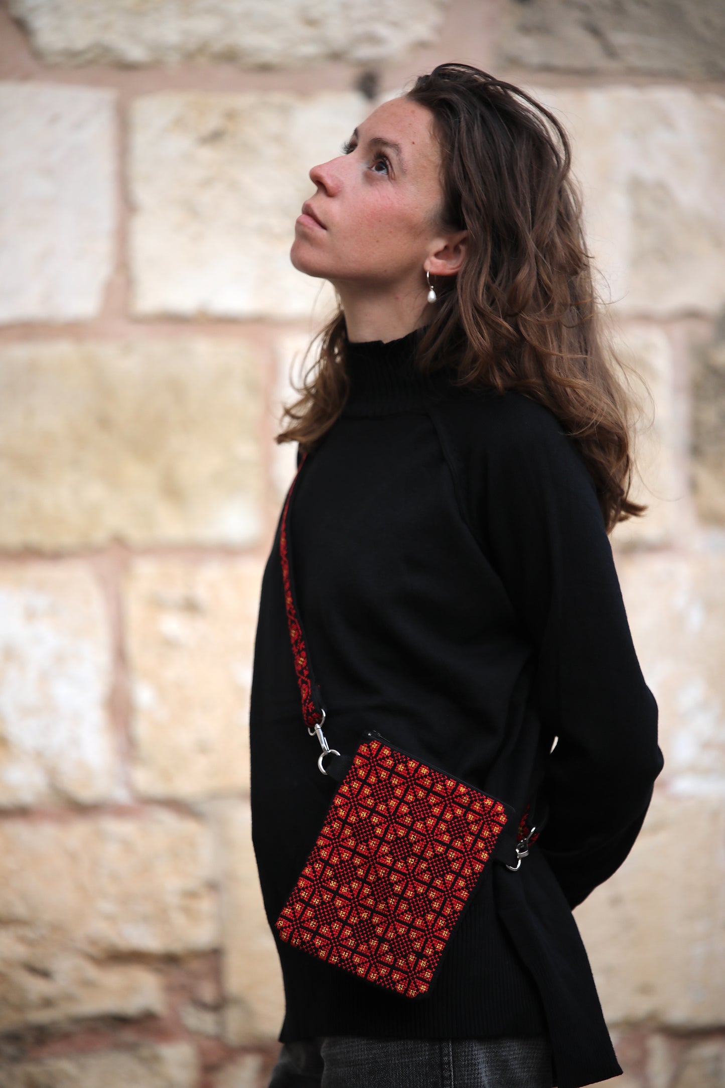 Woman wearing a black coat with a red patterned bag against a stone wall background