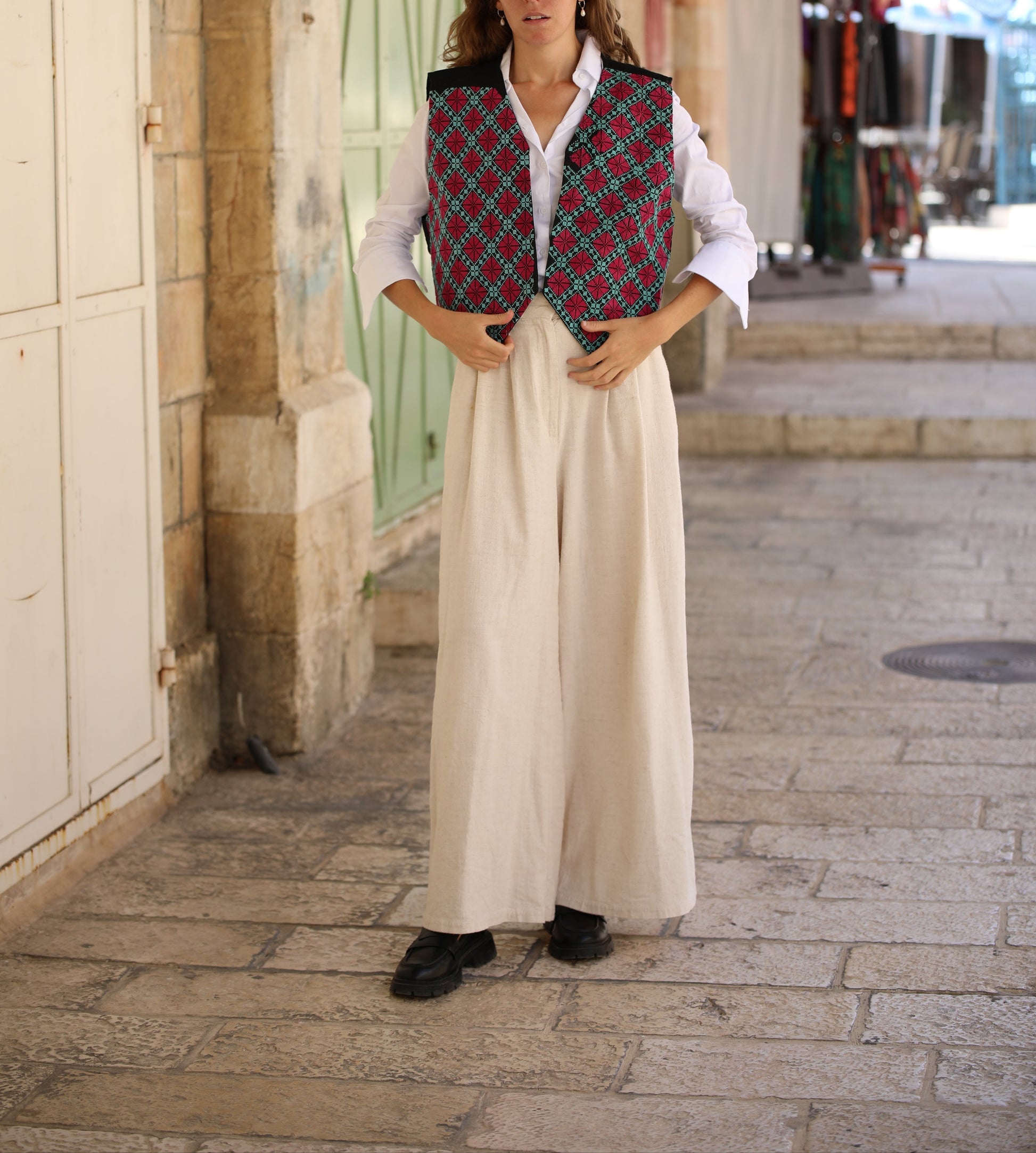 Woman wearing a patterned vest over a white shirt and long beige skirt on a street.