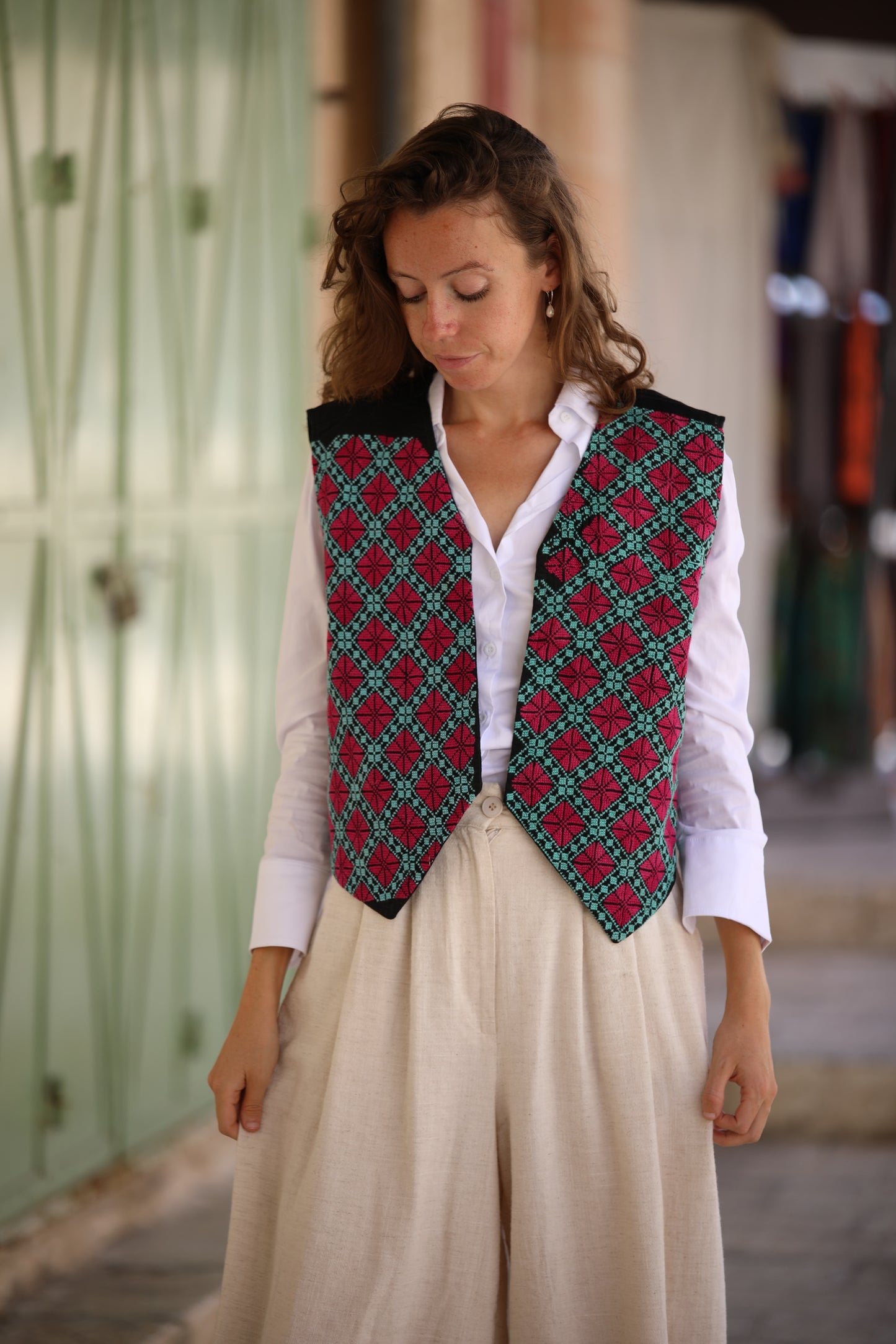 Woman wearing a patterned vest over a white shirt and beige pants in an outdoor setting.