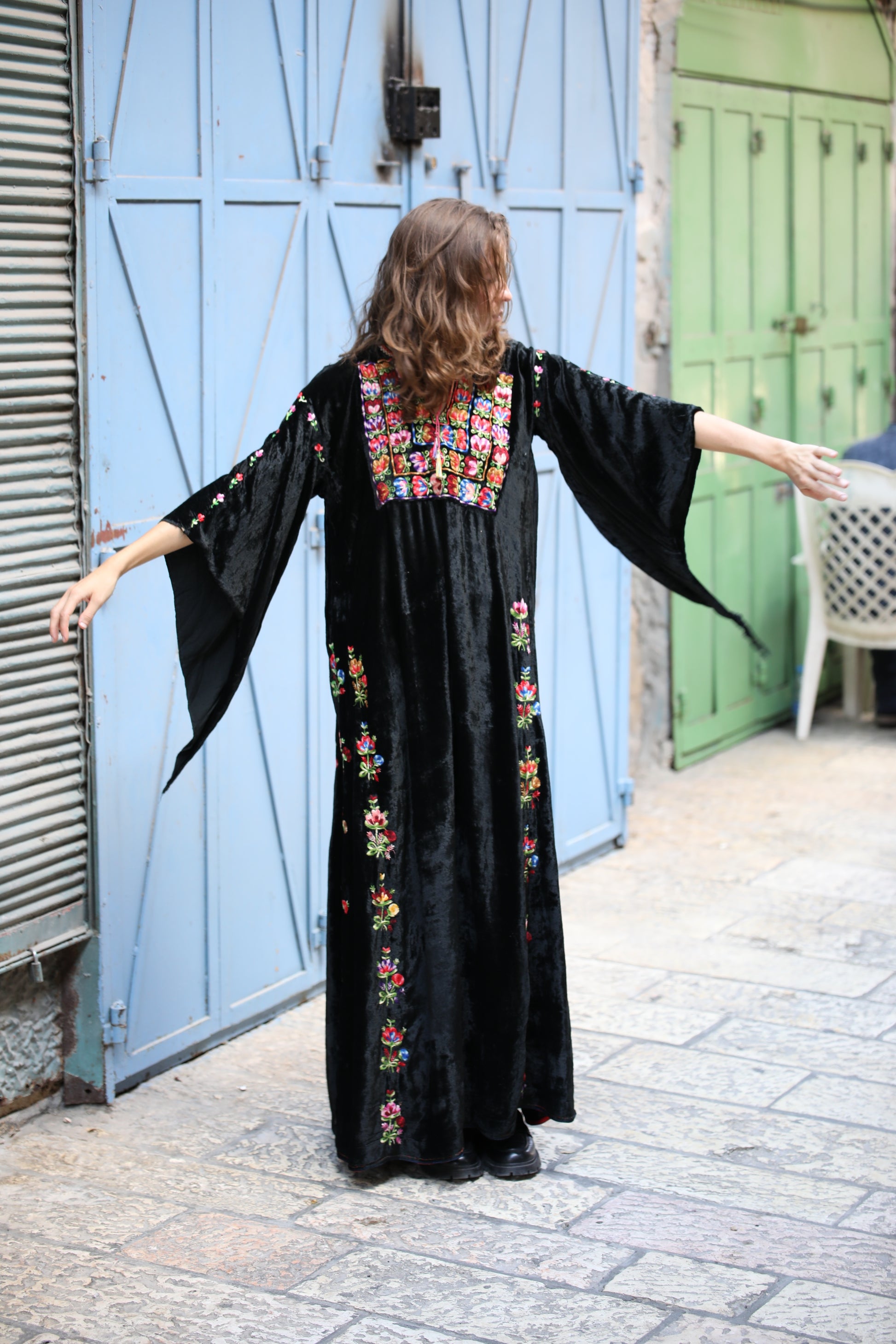 Woman wearing a black dress with colorful embroidery standing in front of blue doors.
