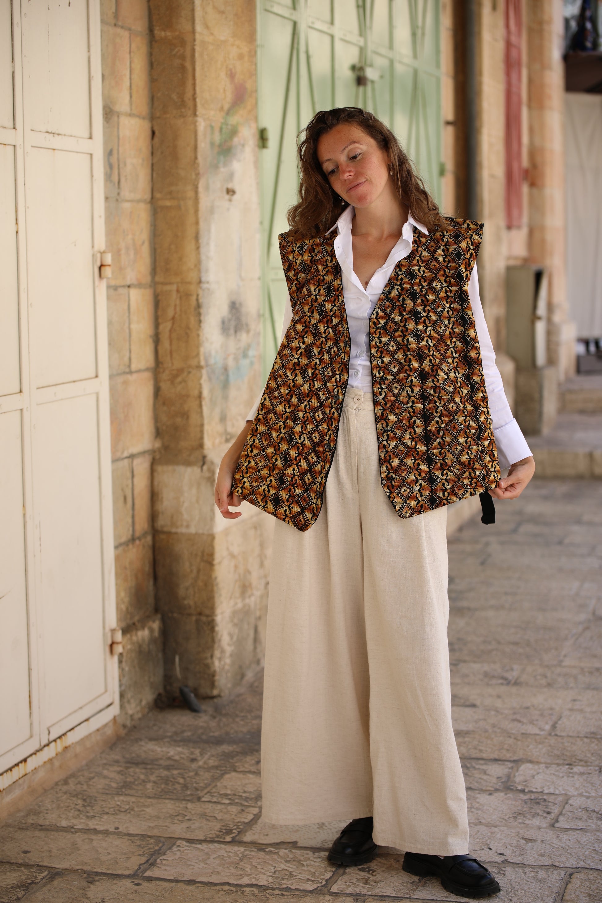 Woman wearing a patterned vest over a white shirt and beige pants, standing on a stone pavement.