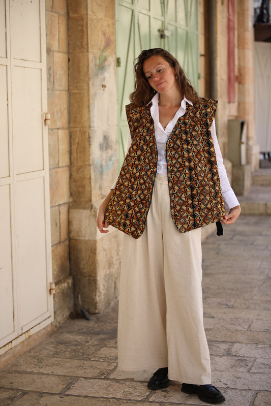 Woman wearing a patterned vest over a white shirt and beige pants, standing on a stone pavement.