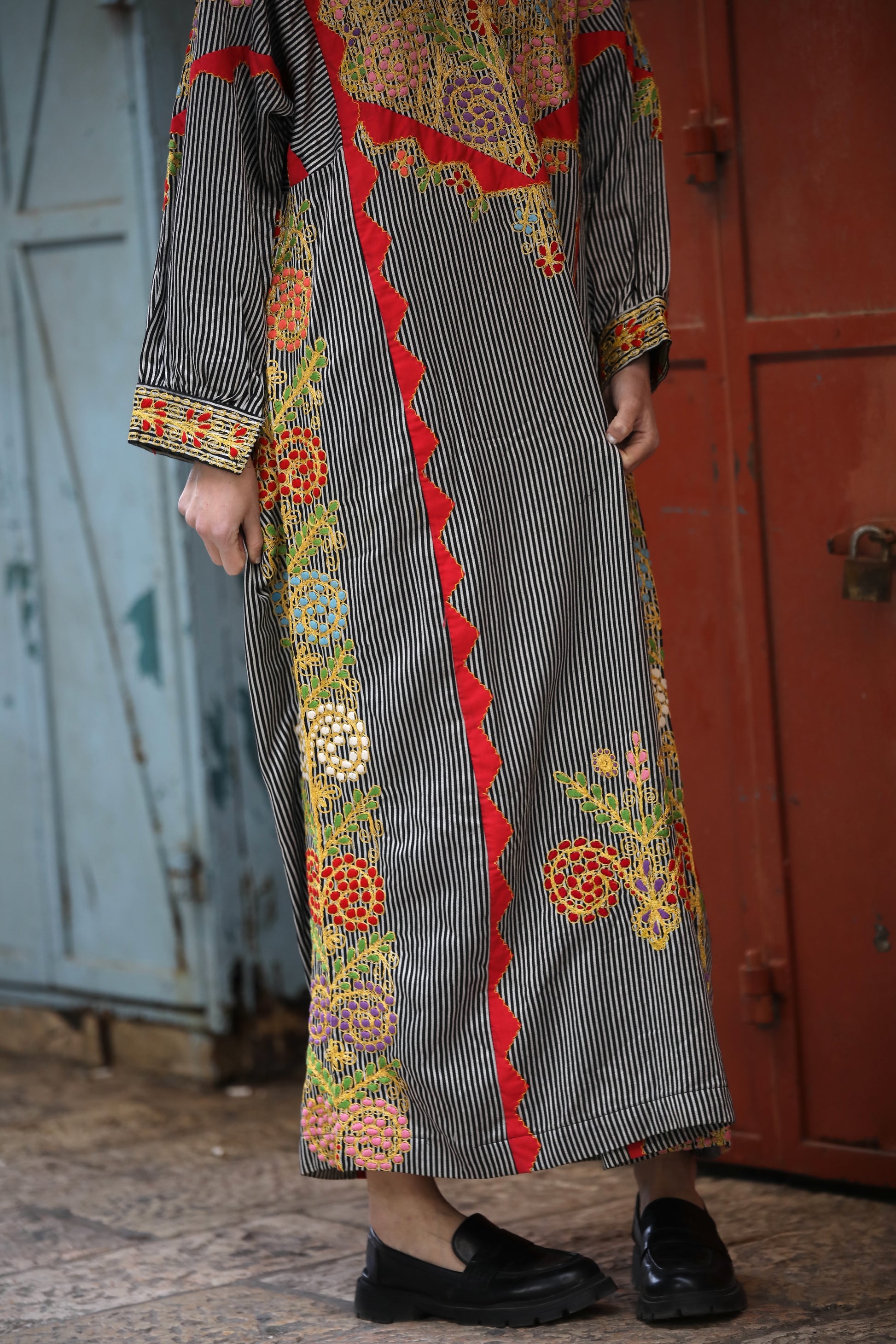 Person wearing a long, embroidered dress with a red border in front of a red door.