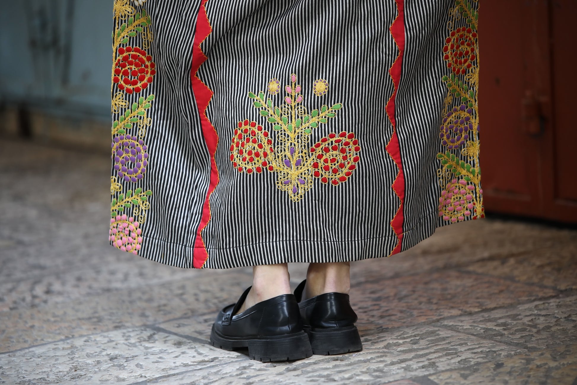 Person wearing a long, embroidered skirt with black shoes on a textured floor.