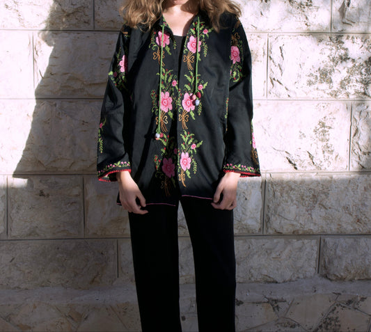 Woman wearing a black embroidered jacket with floral patterns against a stone wall.