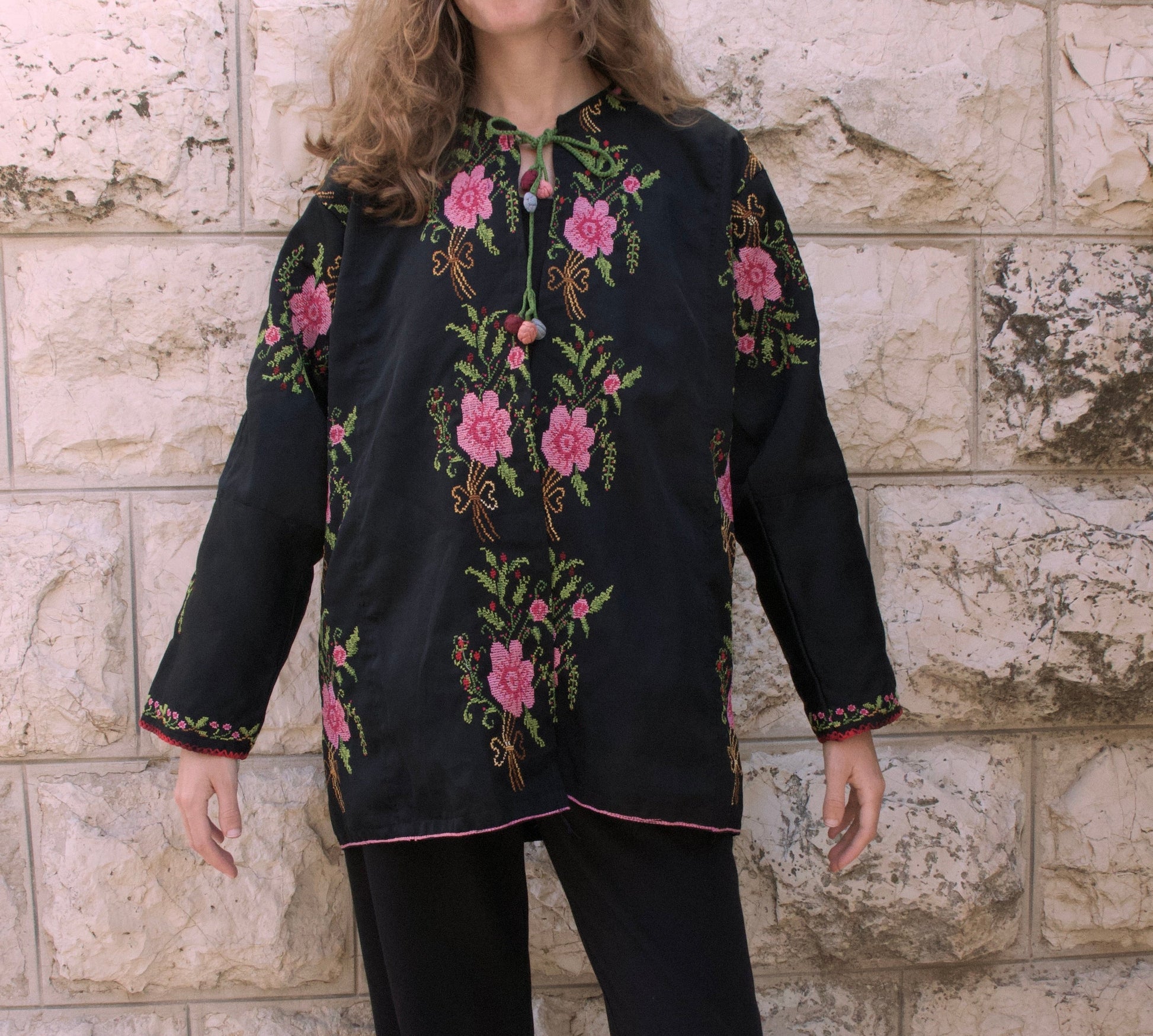 Person wearing a black embroidered shirt and pants standing against a stone wall.