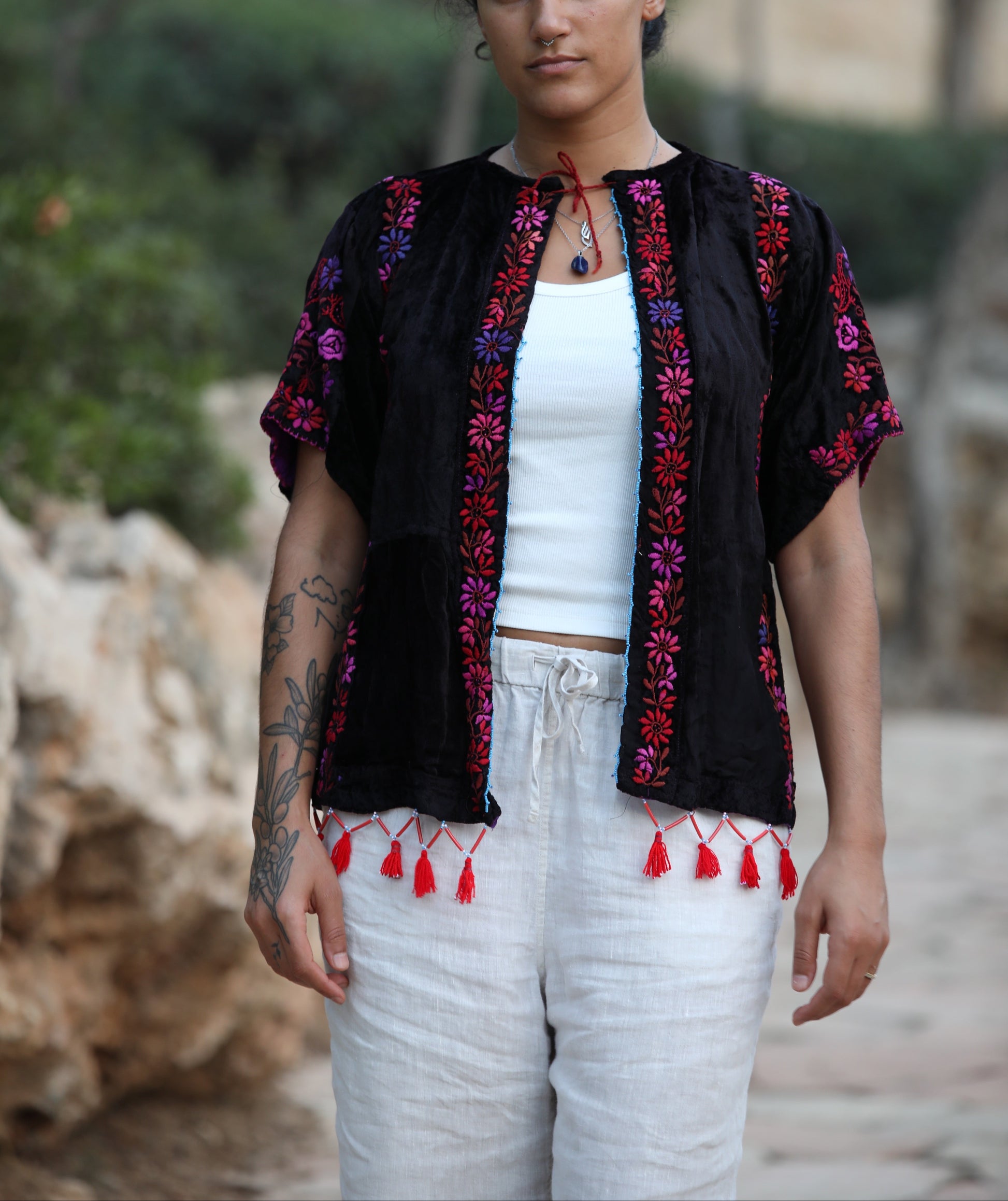 A woman wearing a black embroidered short-sleeve tunic top with red and purple floral patterns and red tassels, styled over a white tank top and white pants, standing outdoors against a blurred natural background.
