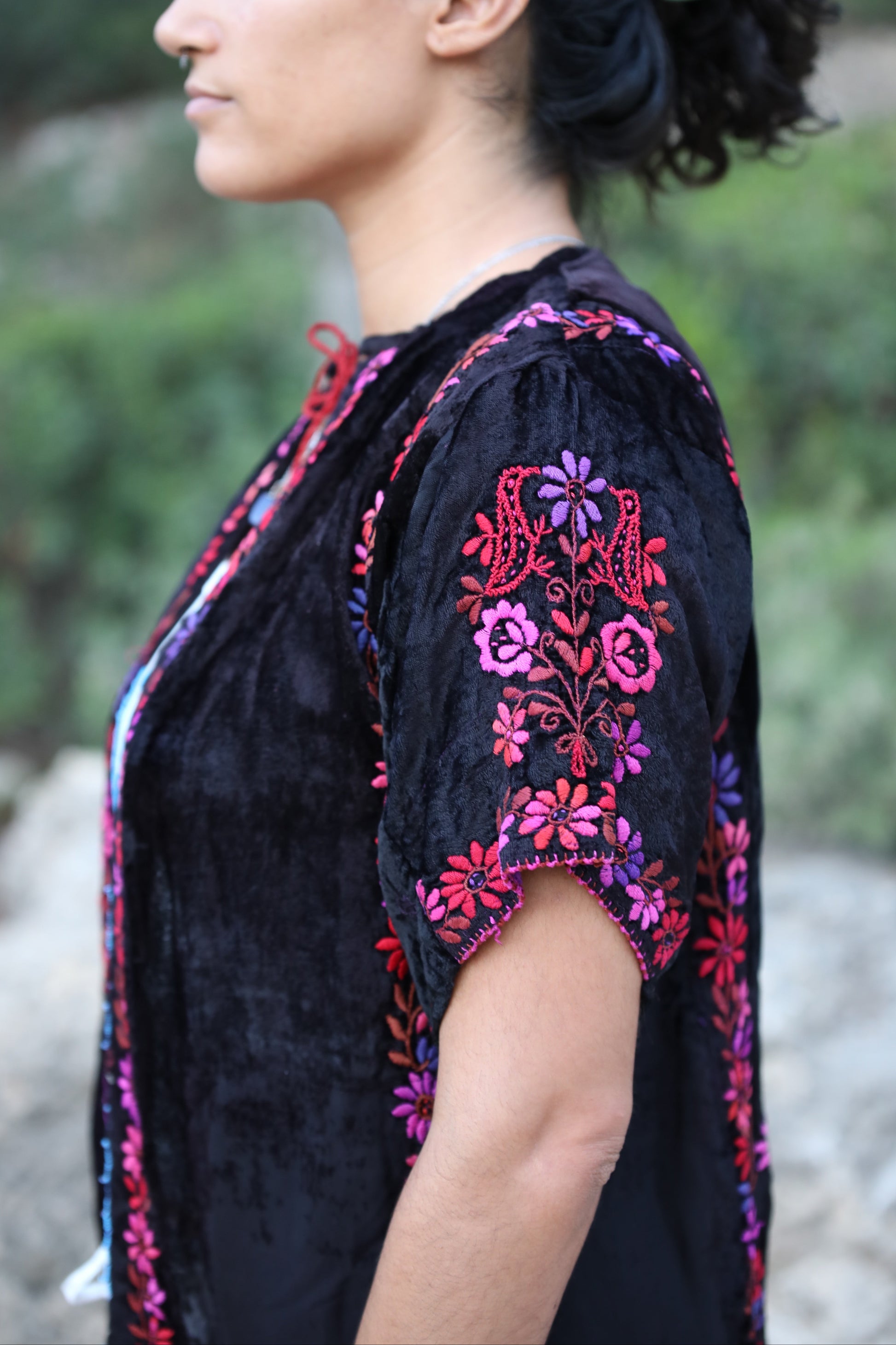A woman shown in profile wearing a black embroidered top with pink, purple, and red floral patterns on the sleeves and neckline, photographed outdoors against a blurred natural background.