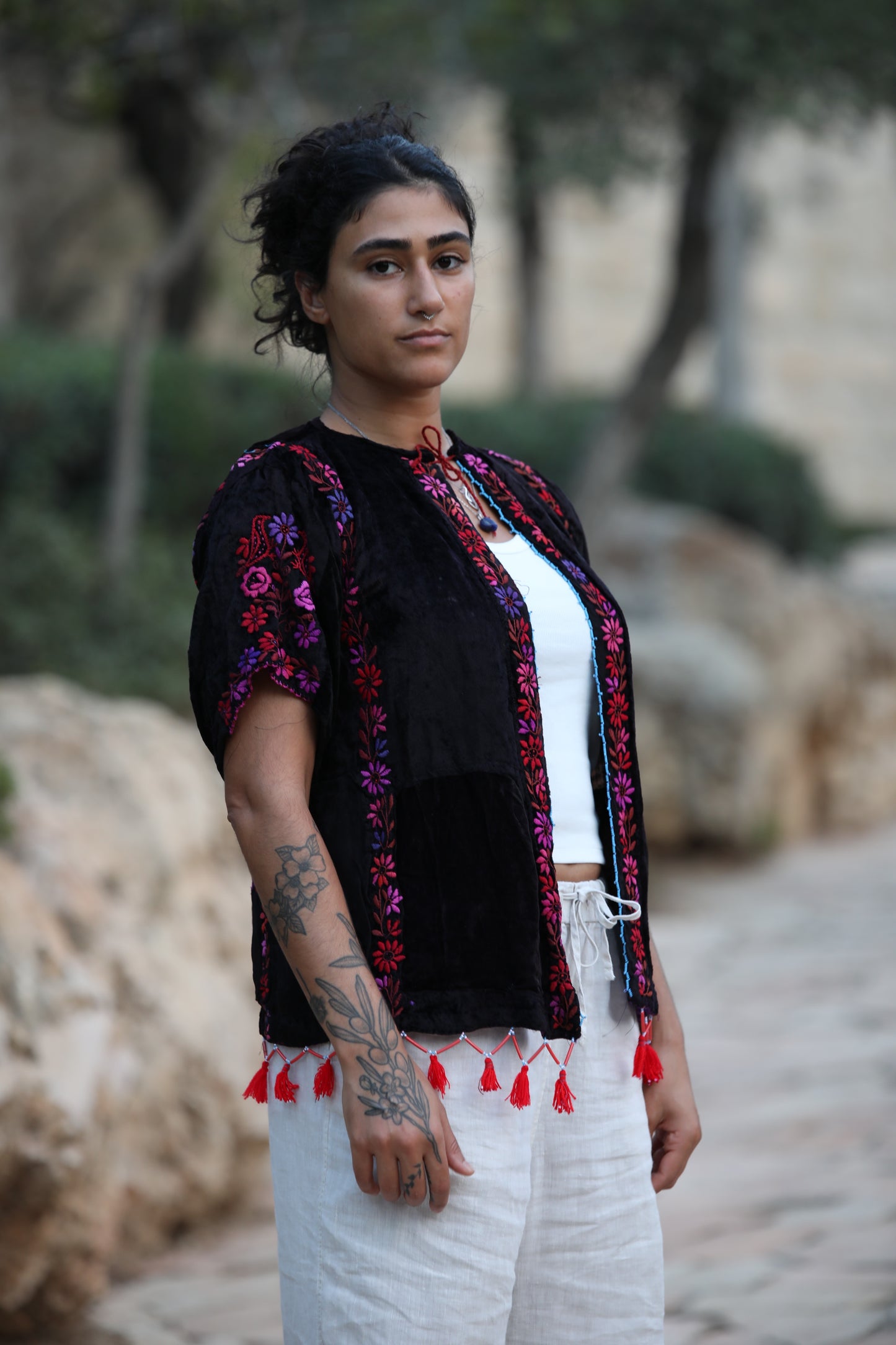 A woman wearing a black embroidered short-sleeve jacket with red tassels over a white top and white pants, standing outdoors on a stone path with trees and rocks in the background.