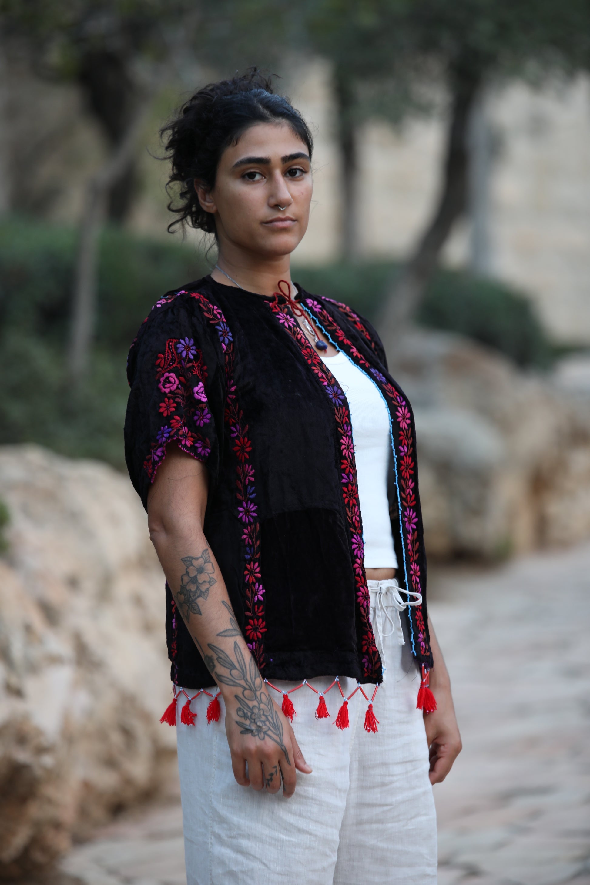 A woman wearing a black embroidered short-sleeve jacket with red tassels over a white top and white pants, standing outdoors on a stone path with trees and rocks in the background.