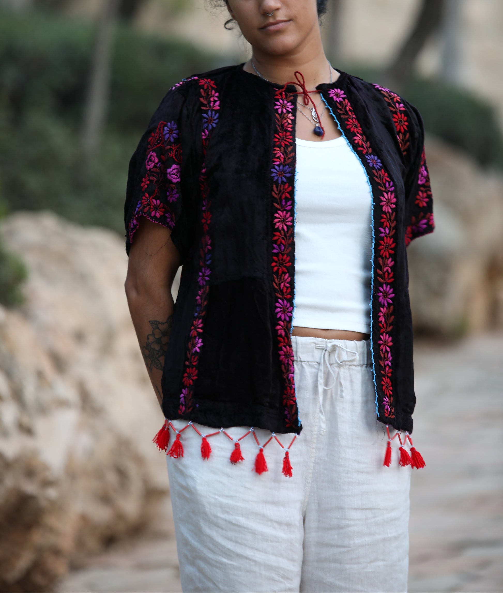 A woman wearing a black embroidered short-sleeve blouse with red and purple floral patterns and red tassels, paired with a white tank top and white pants, standing outdoors against a blurred natural background.