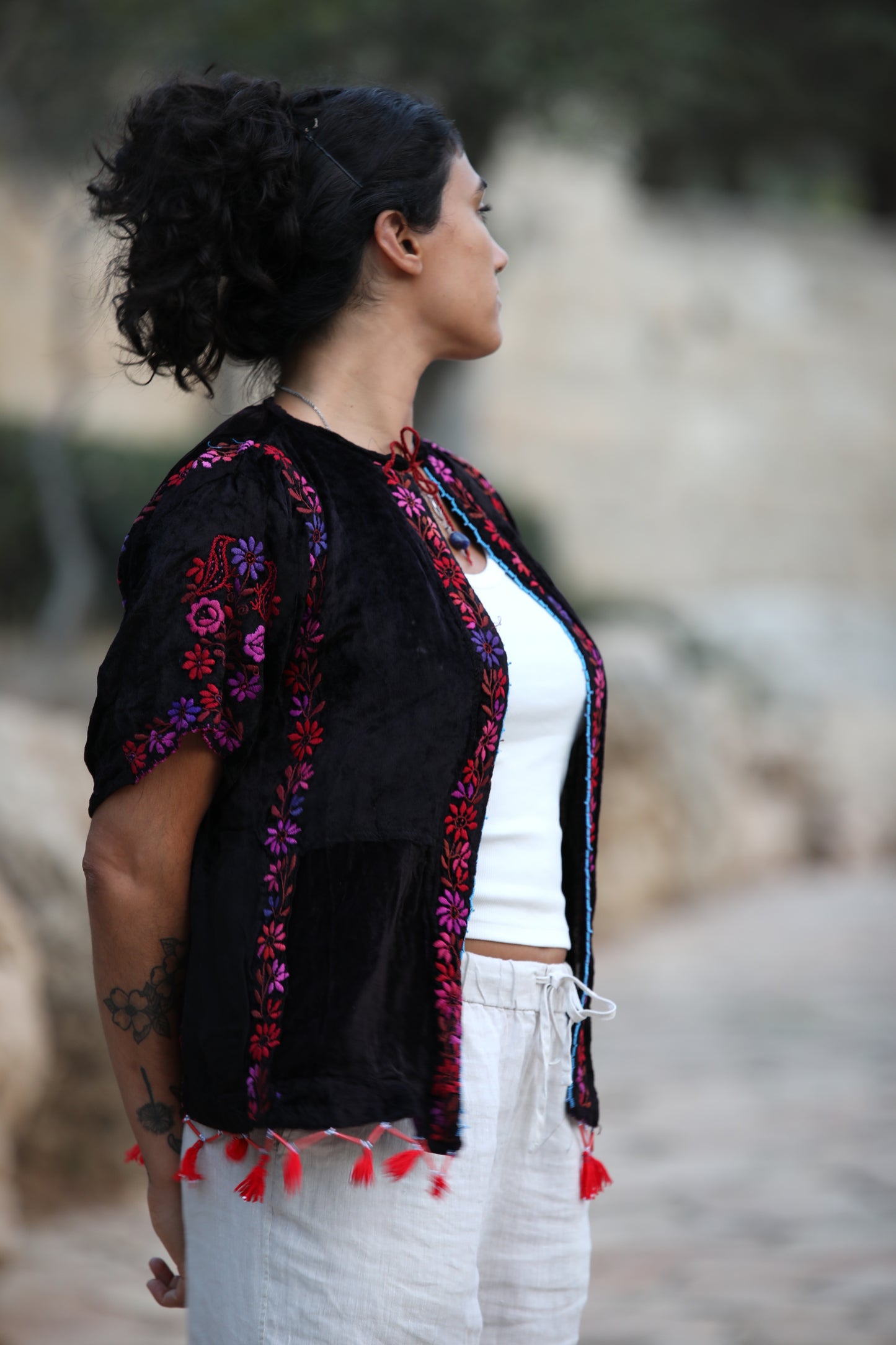 A woman shown from the side in an outdoor setting, wearing a black embroidered cardigan with colorful floral patterns and red tassels over a white top and white pants.