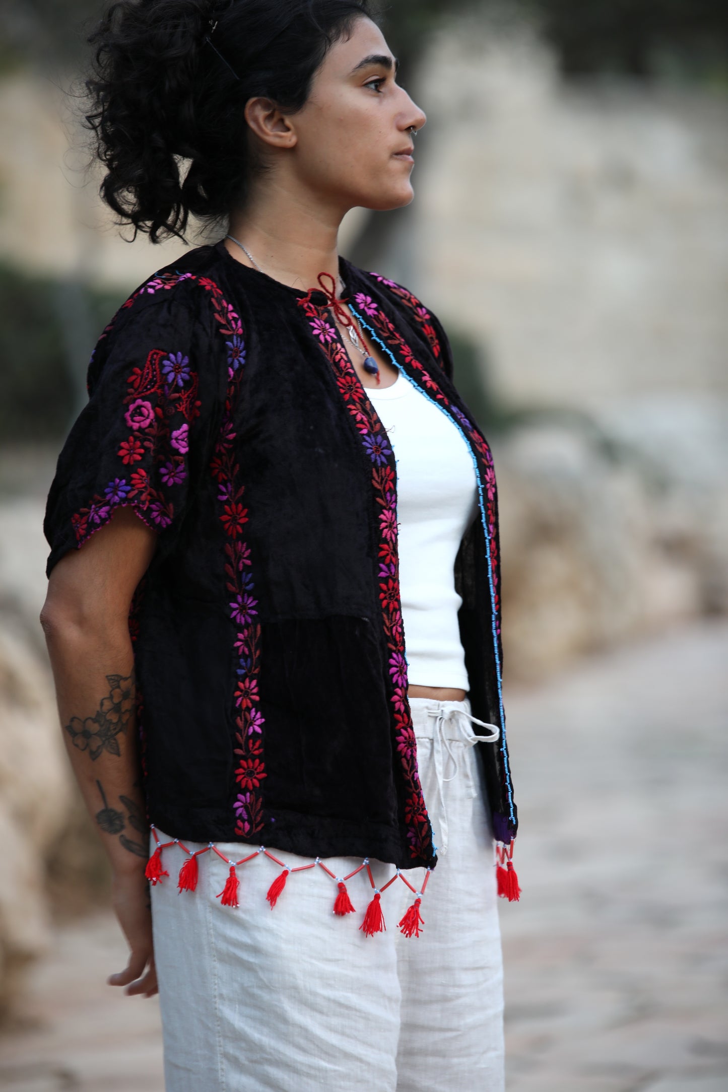 A woman wearing a black embroidered traditional-style jacket with colorful floral patterns and red tassels, layered over a white top and white pants, photographed outdoors against a blurred stone background.