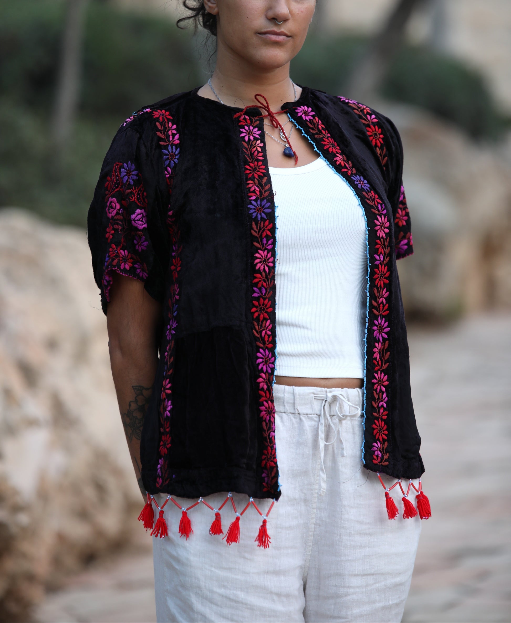 A woman wearing a black embroidered short-sleeve jacket with red, purple, and pink floral patterns and red tassels, styled over a white tank top and white pants, standing outdoors against a blurred rocky background.