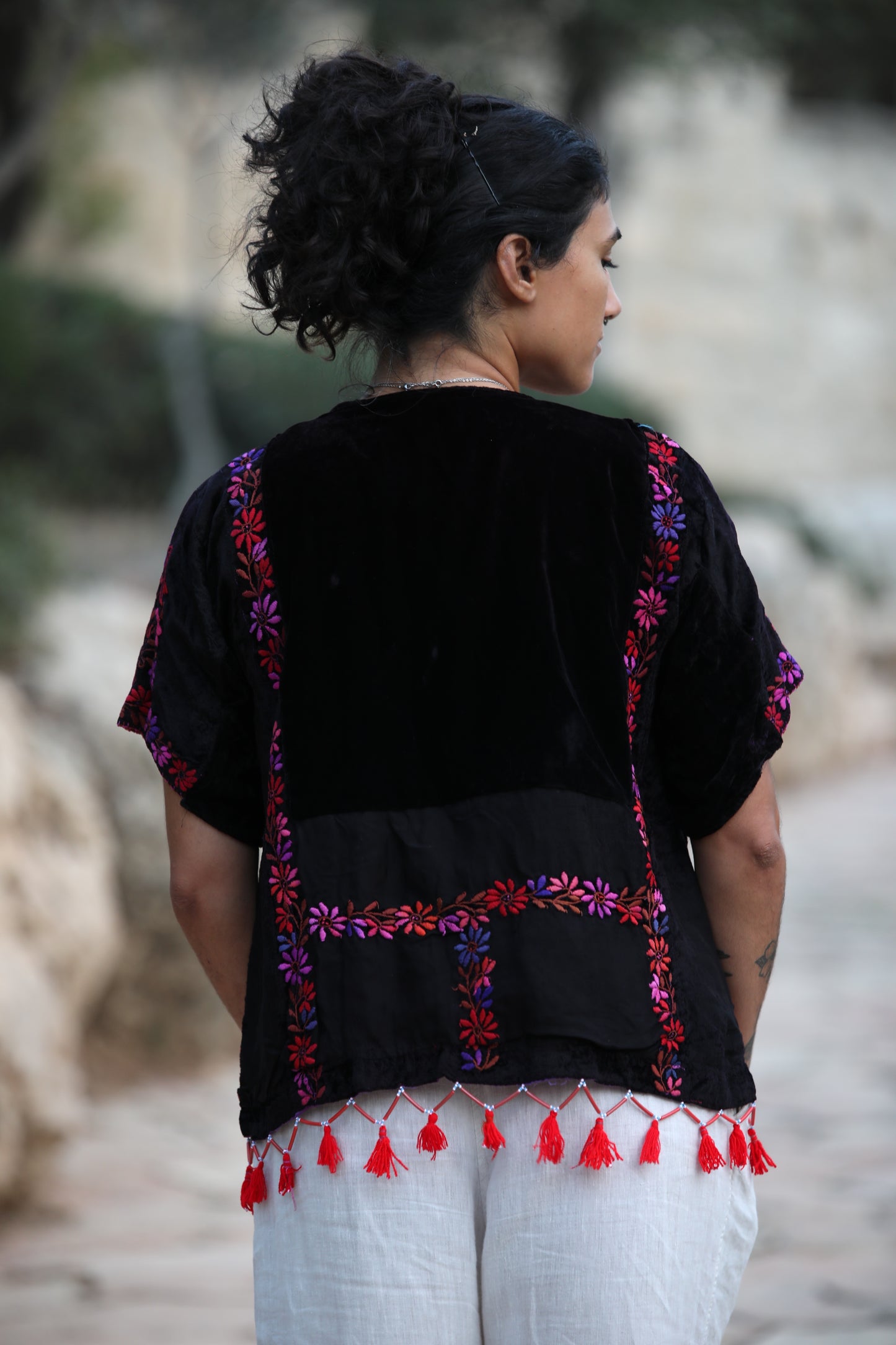 A woman viewed from behind wearing a black embroidered blouse with colorful floral embroidery and red tassels, paired with white pants, in an outdoor setting.