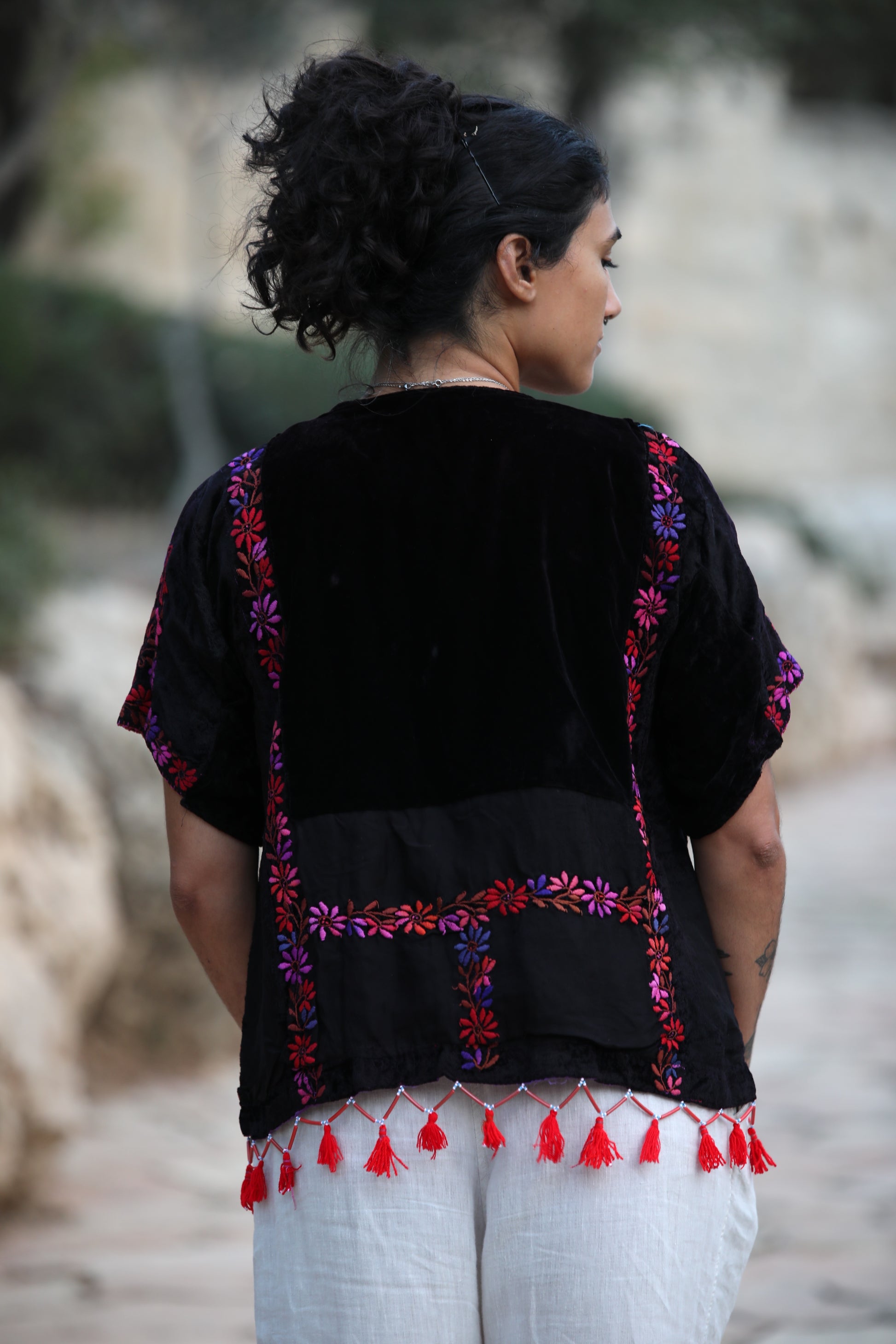 A woman viewed from behind wearing a black embroidered blouse with colorful floral embroidery and red tassels, paired with white pants, in an outdoor setting.