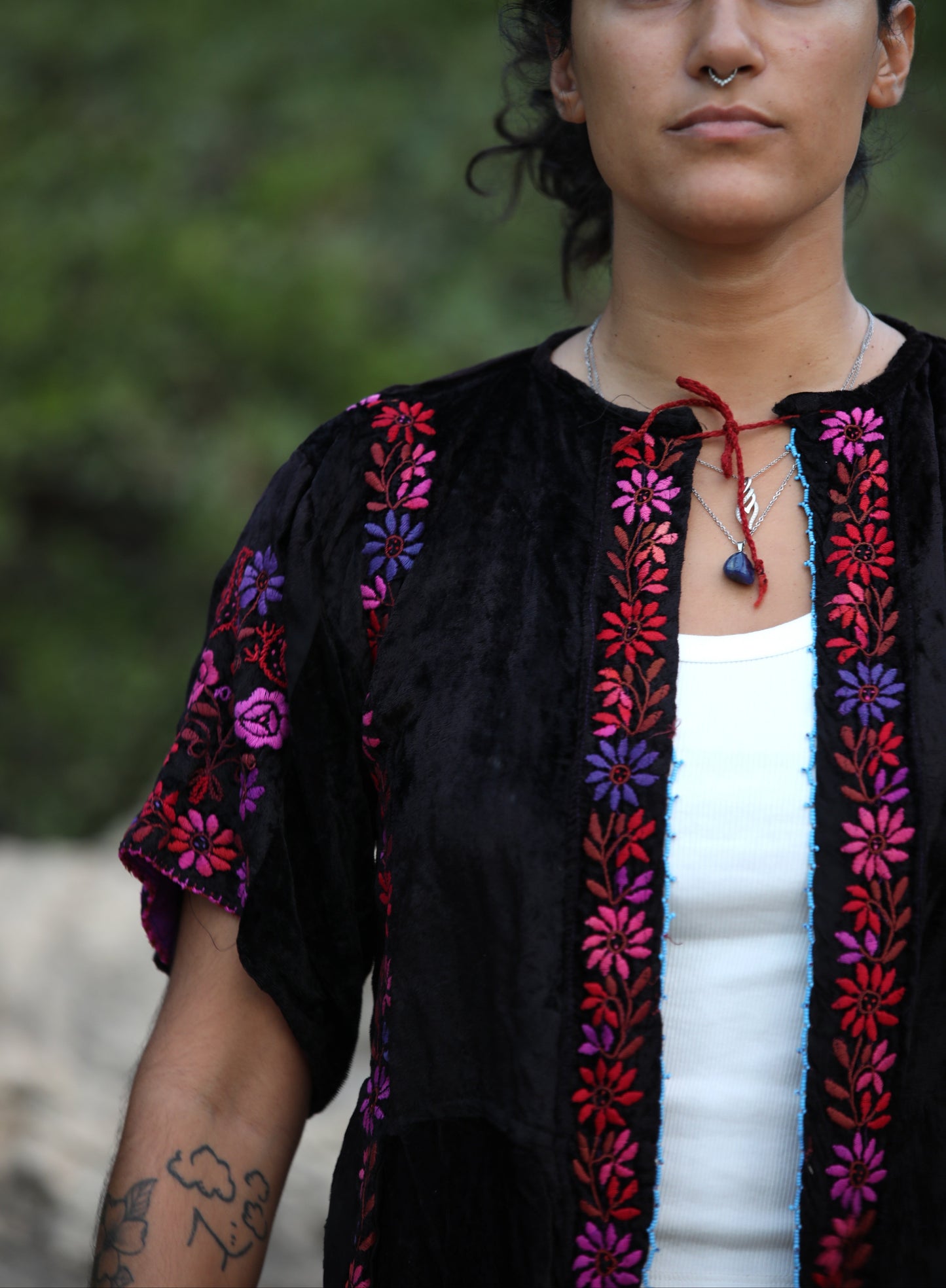 A woman wearing a black velvet embroidered blouse with vibrant pink, purple, and red floral patterns, layered over a white tank top, set against a blurred green outdoor background.