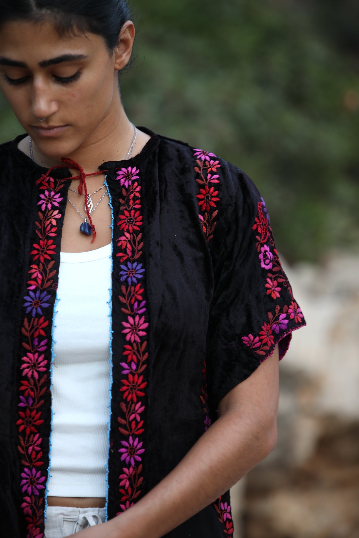 A close-up of a woman wearing a black velvet short-sleeve cardigan with vibrant pink, purple, and red floral embroidery, layered over a white tank top.