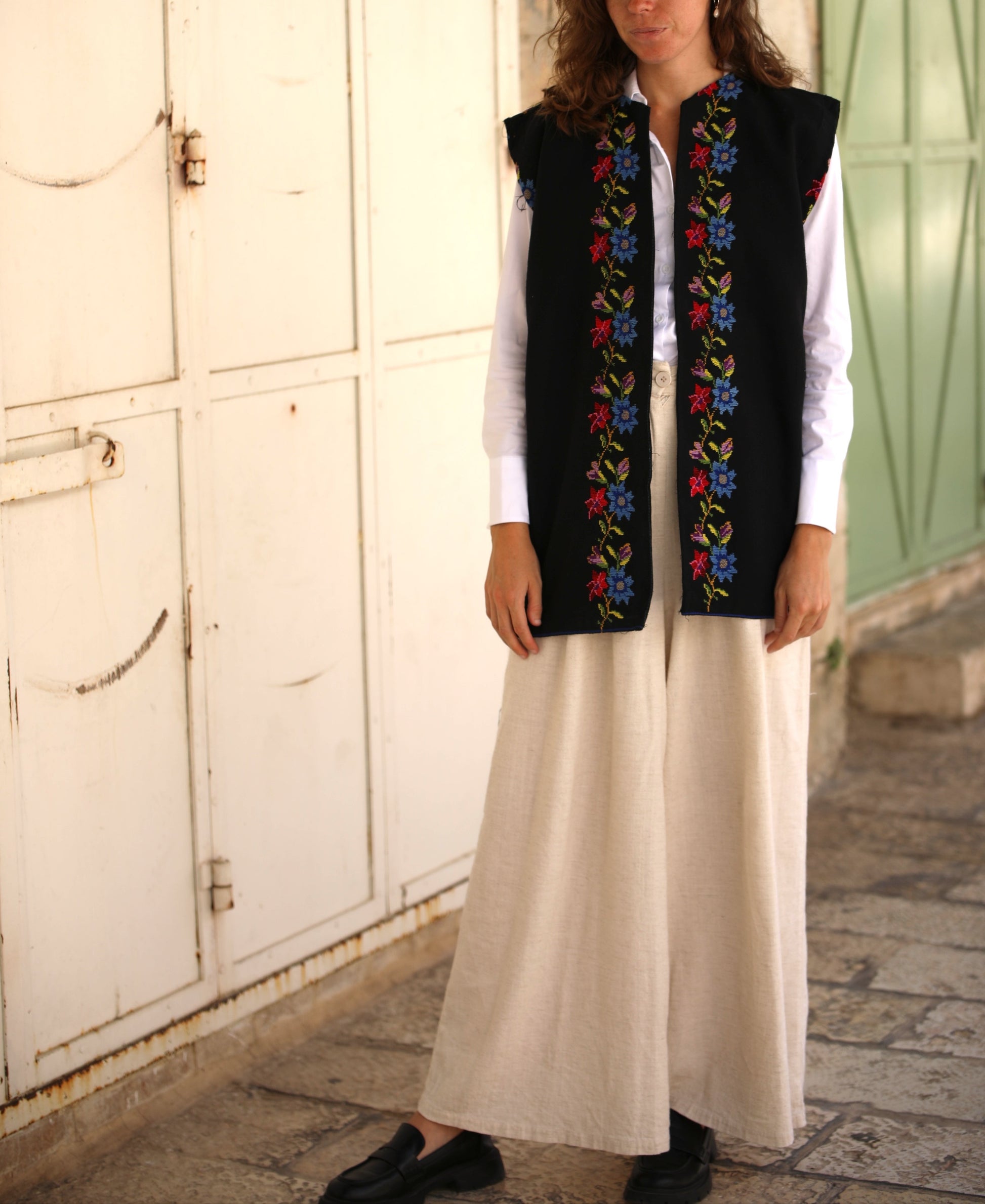 Woman wearing a black embroidered vest over a white shirt and beige pants, standing in front of a white wall.