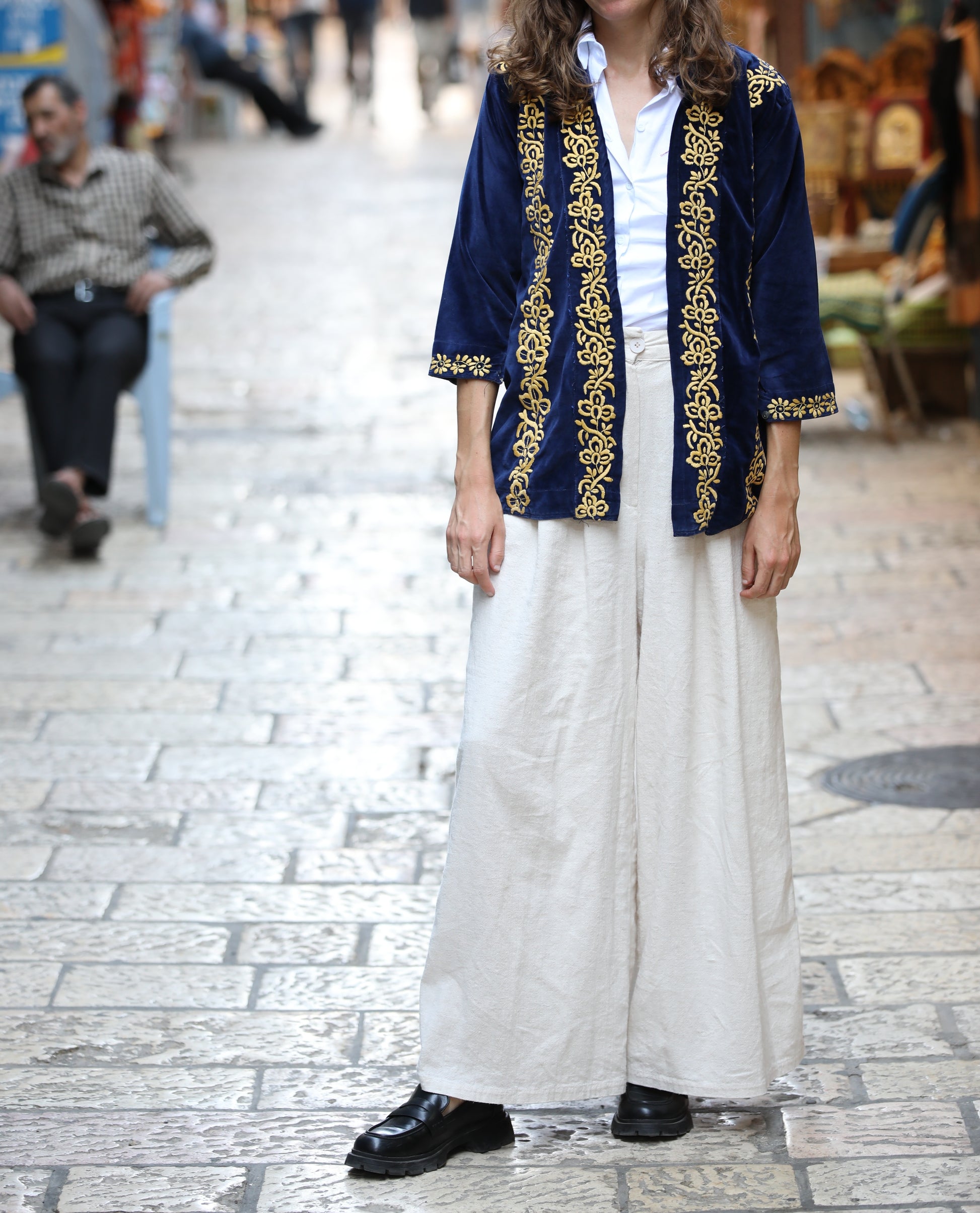 Person wearing a blue embroidered jacket and white pants on a stone pavement.