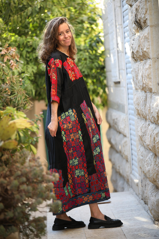 Woman wearing a colorful embroidered dress standing outdoors near a stone wall.