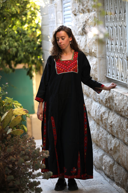 Woman wearing a black dress with red patterns standing against a stone wall.