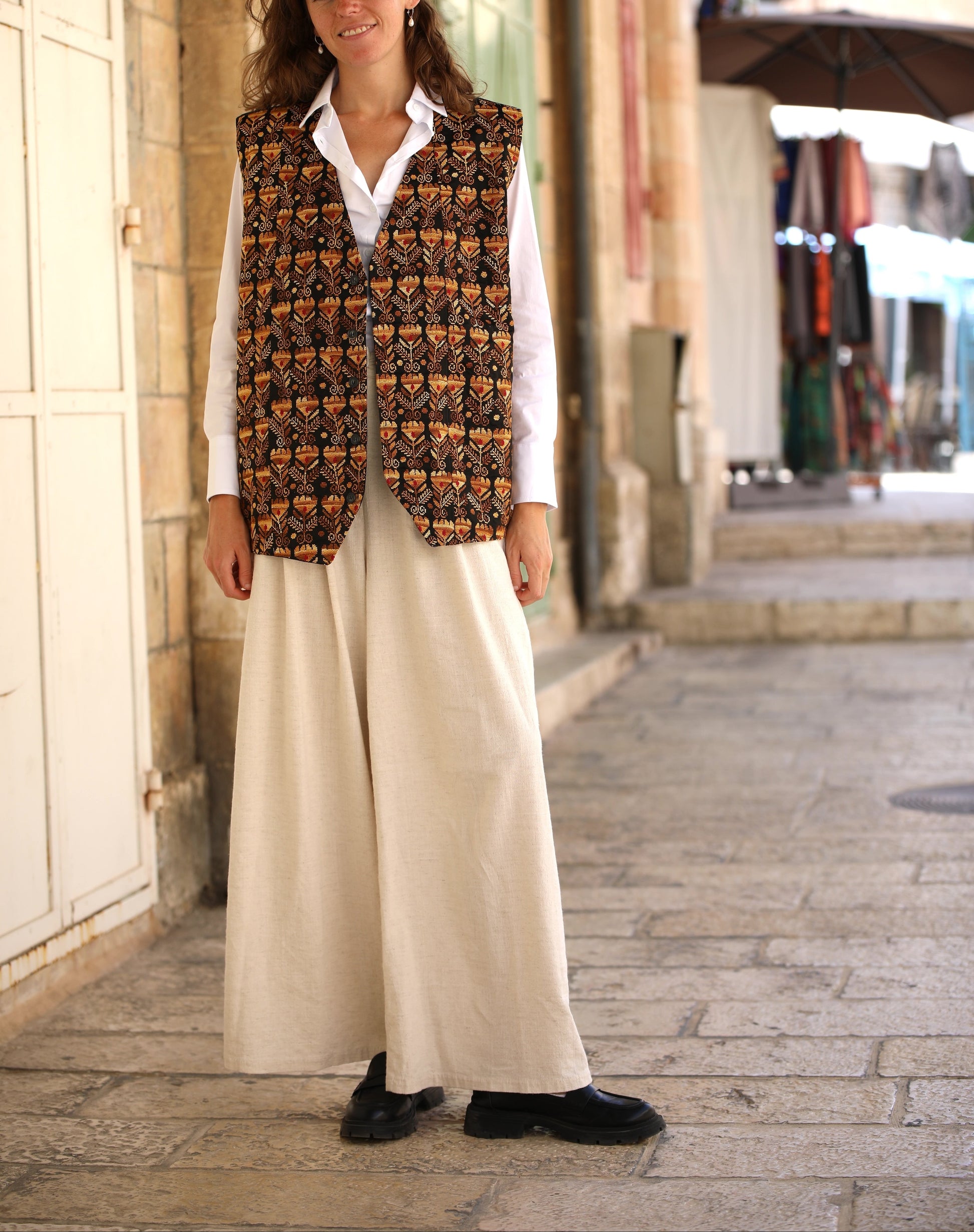 Woman wearing a patterned vest and white pants standing on a street.