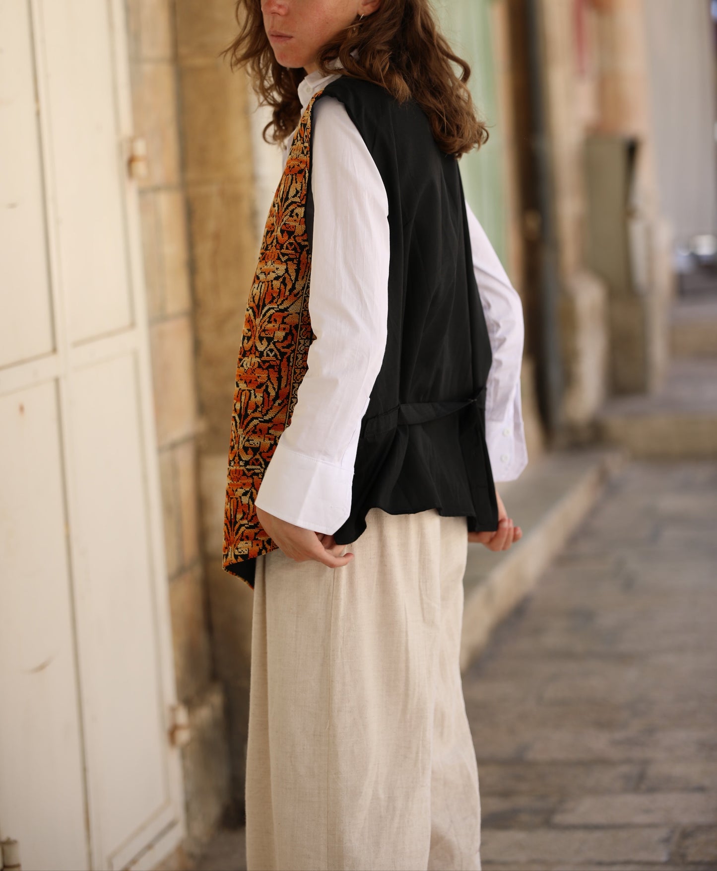 Woman standing in a narrow alleyway wearing a white shirt, black vest, and patterned scarf.