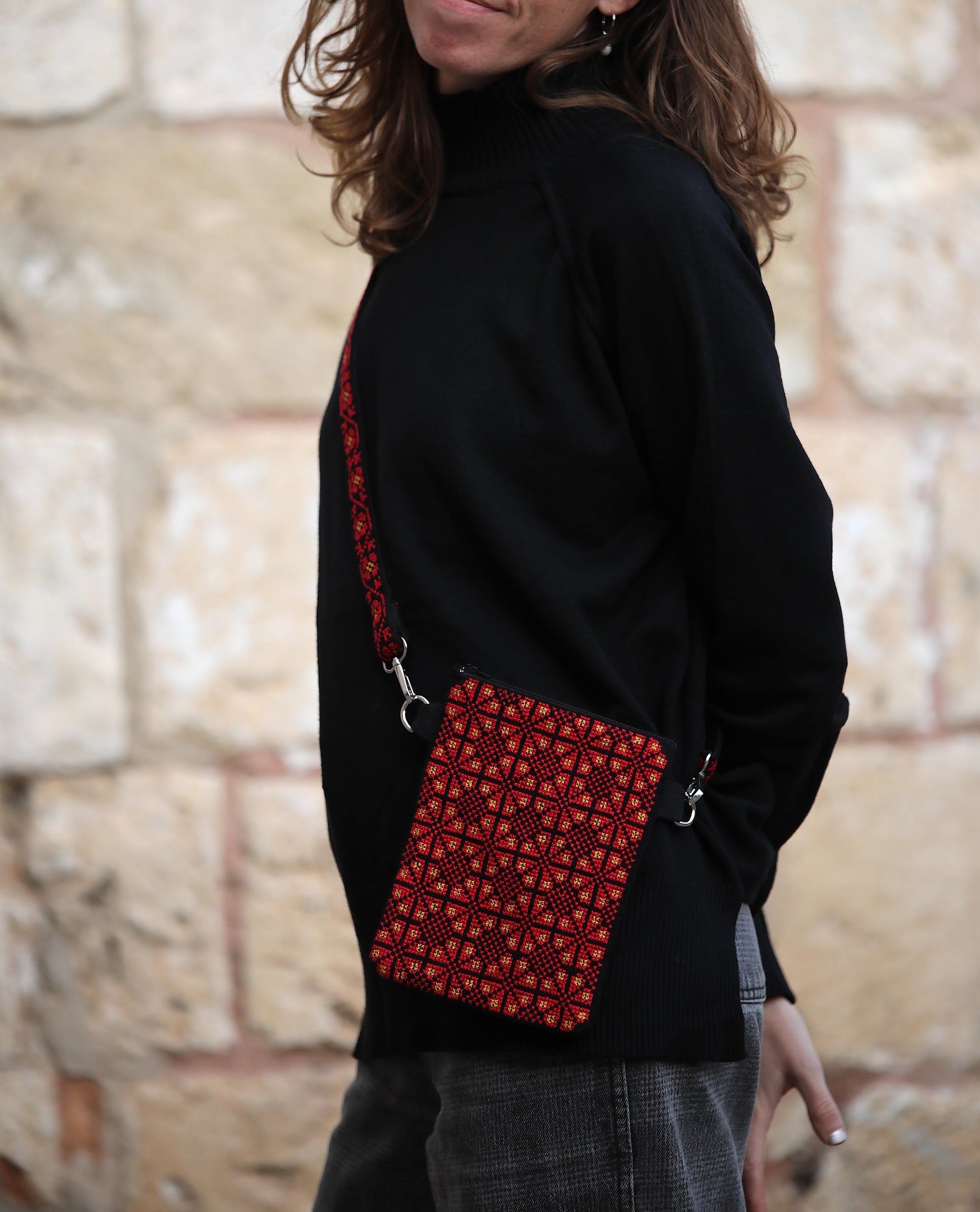 Person wearing a black sweater with a red patterned bag over their shoulder against a stone wall background.