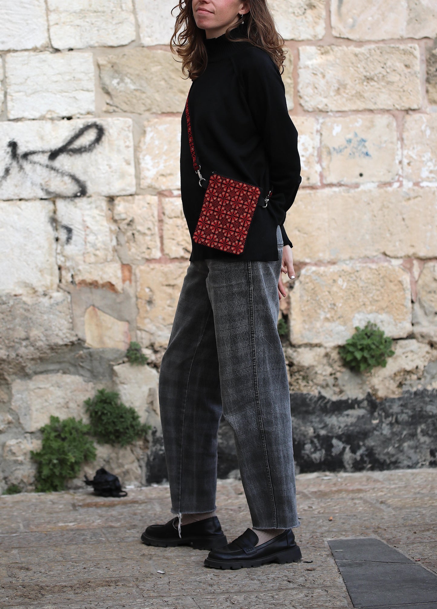 Person wearing a black sweater, plaid pants, and carrying a red patterned bag against a stone wall.
