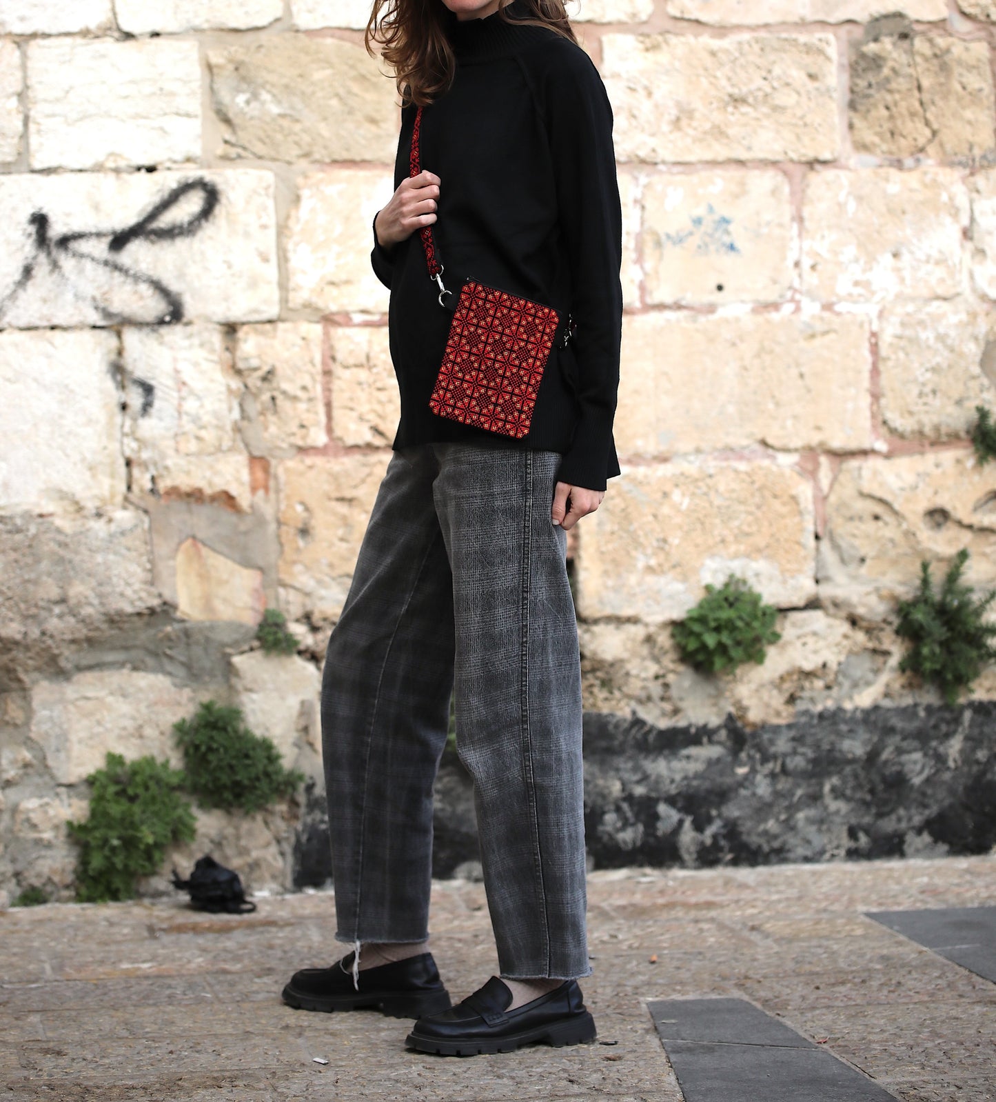 Person wearing a black sweater and gray pants holding a red patterned bag against a stone wall background.