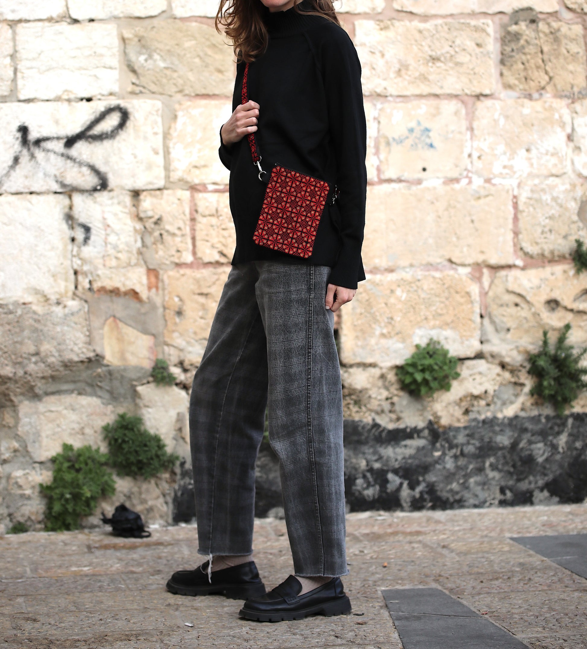 Person wearing a black sweater and gray pants holding a red patterned bag against a stone wall background.