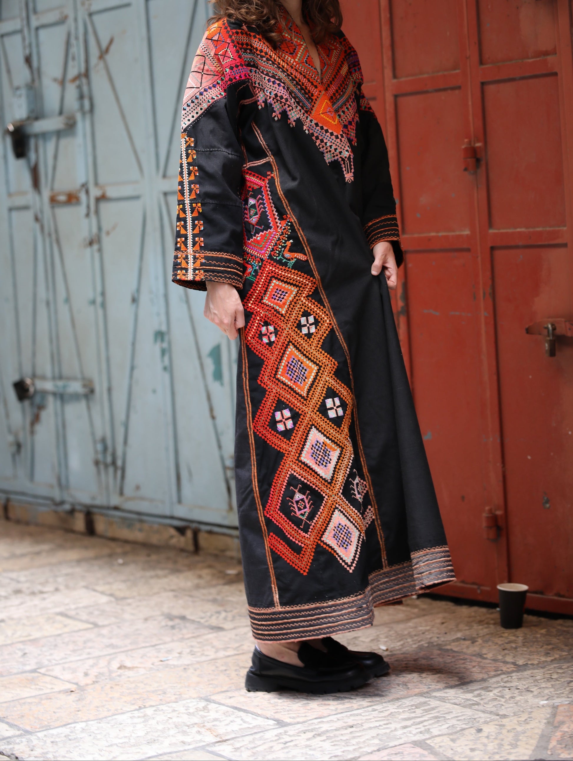 Woman wearing a long, embroidered dress with geometric patterns in front of a textured wall.