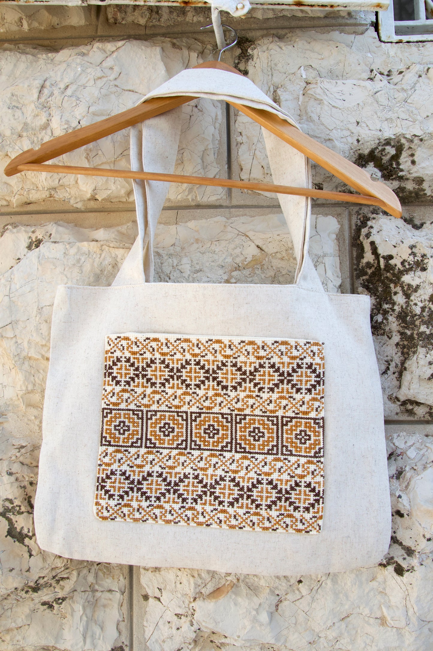 White tote bag with decorative pattern on a wooden hanger against a stone wall.