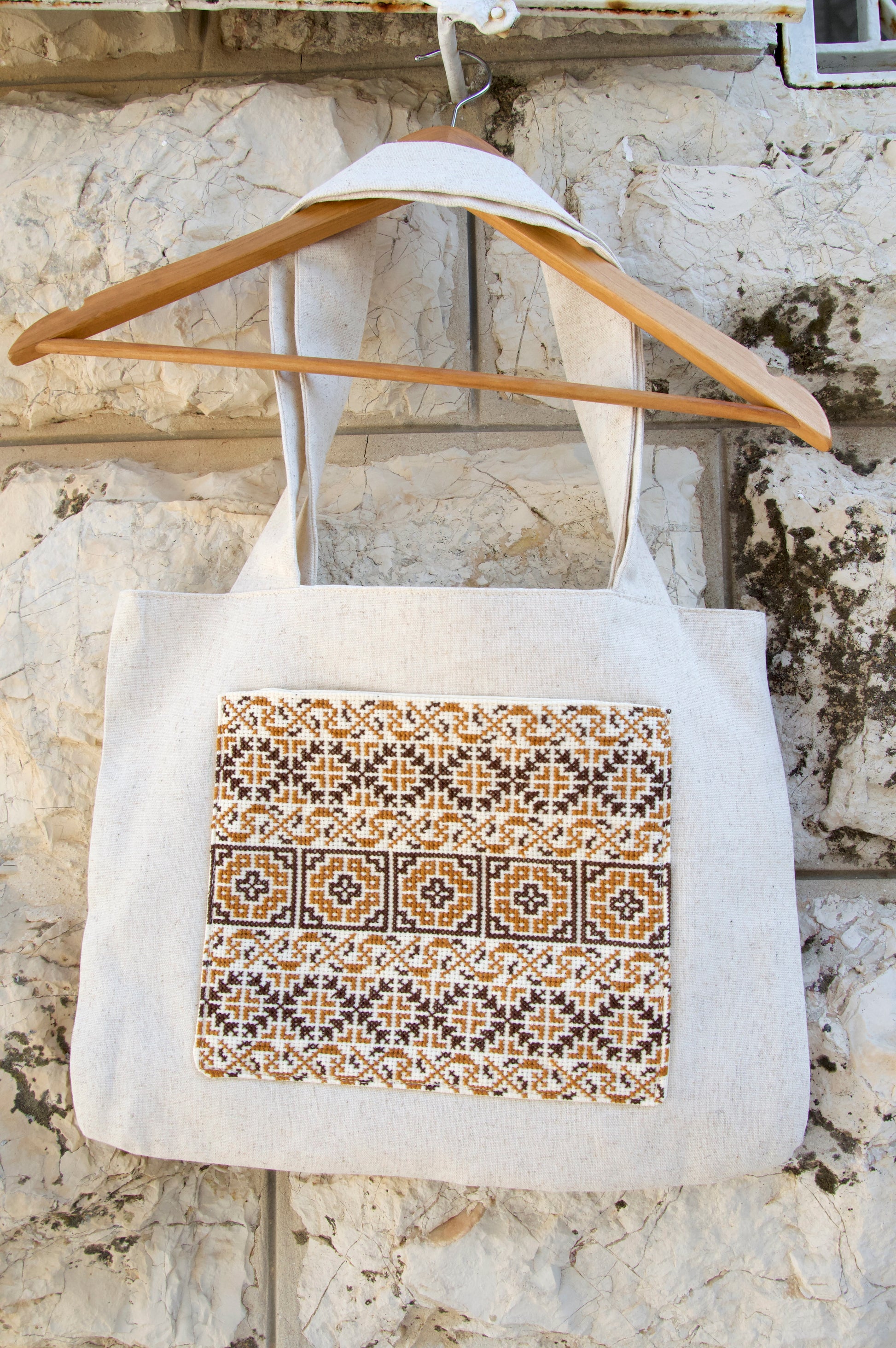 White tote bag with decorative pattern on a wooden hanger against a stone wall.