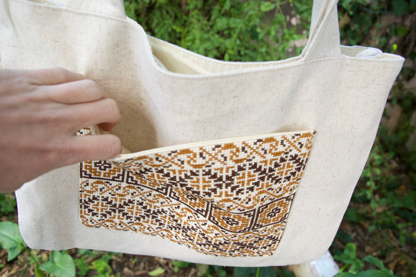 Beige tote bag with a patterned pocket held by a hand against a blurred green background
