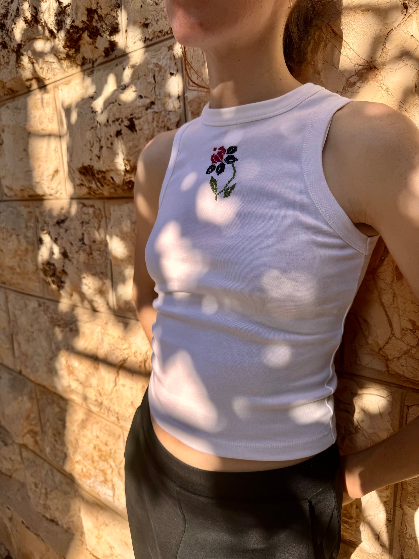 Person wearing a white sleeveless top with floral embroidery against a stone wall.