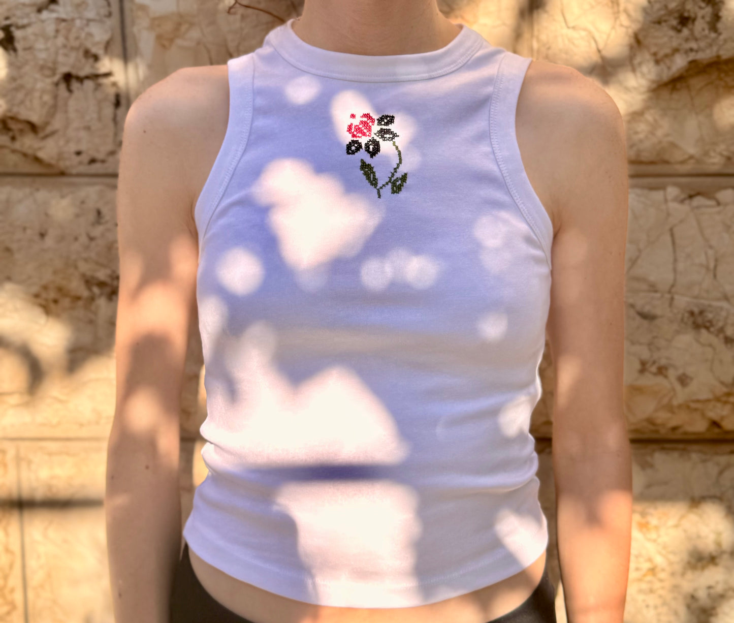 Person wearing a white tank top with floral embroidery against a stone wall.
