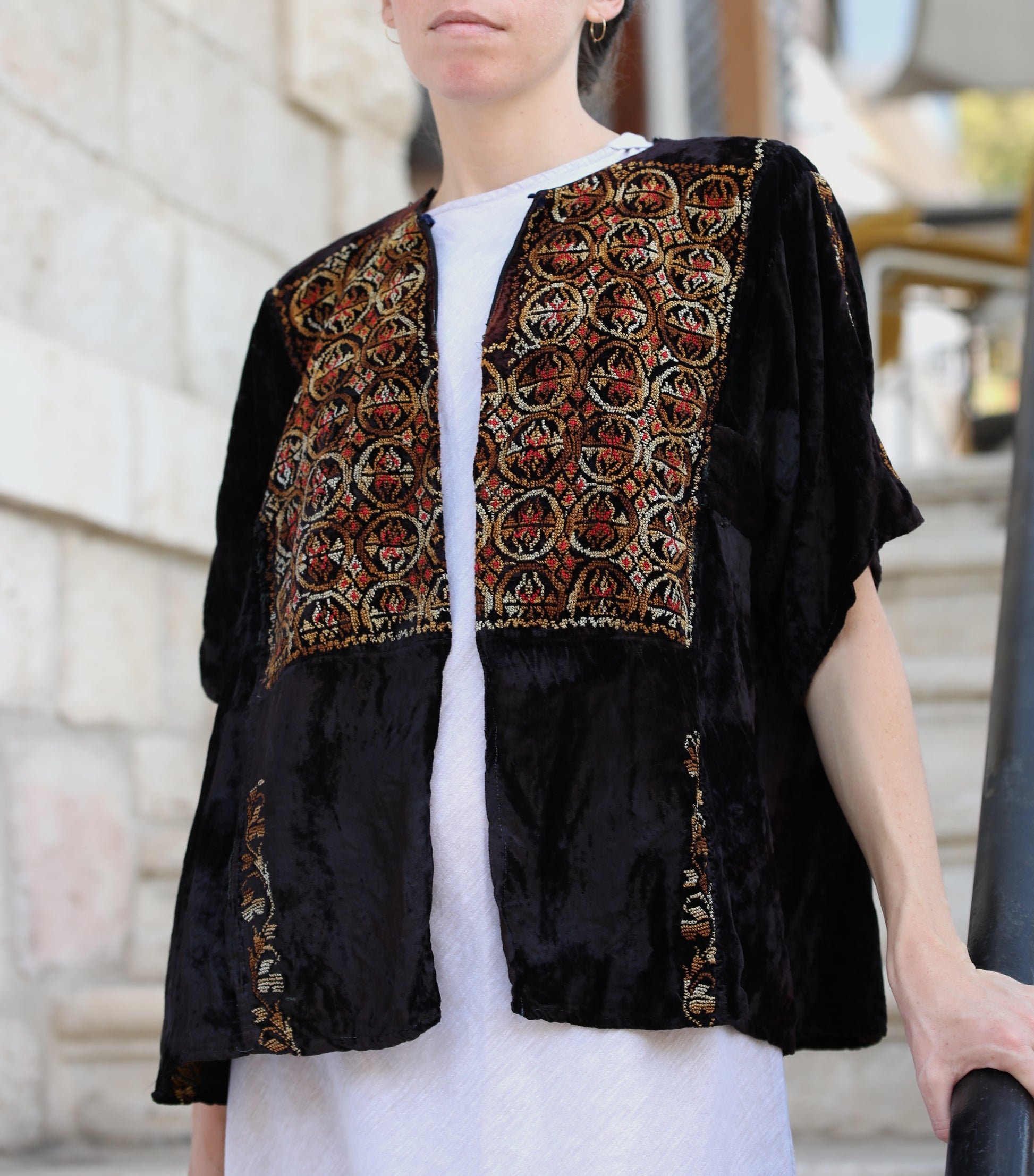 A person wearing a black velvet short-sleeve jacket with ornate multicolor embroidered panels on the front, styled over a white top in an outdoor setting.